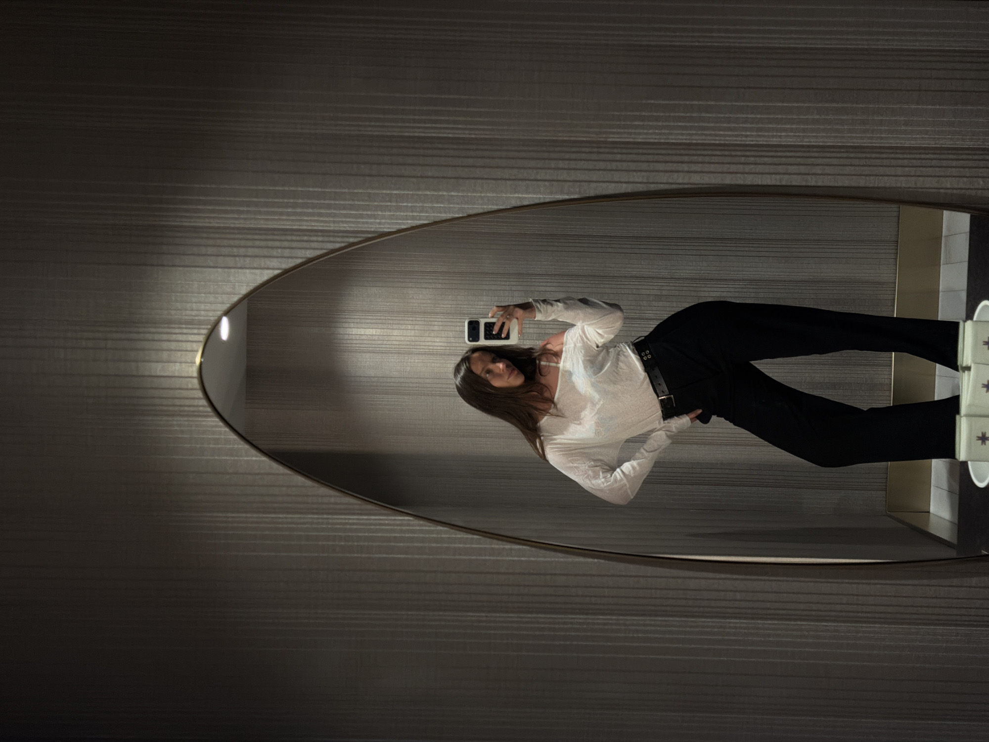 This image is an indoor mirror selfie taken by a young adult female, likely within a hotel or private residence in Budva, Montenegro. The central subject, a woman with long, dark hair, is reflected from the waist up in a large, floor-length, irregularly shaped oval mirror with a thin, golden-bronze frame. She is wearing a white, off-the-shoulder long-sleeved top and high-waisted black trousers secured with a black belt. Her left hand holds a white smartphone, positioned to capture her reflection, while her right hand rests on her hip. She is posed with her body angled slightly, looking towards the phone's camera. The wall behind the mirror is covered with dark gray or taupe textured wallpaper featuring subtle vertical stripes. A distinct bright light source is reflected at the top of the mirror's arch. At the bottom of the reflection, two small, rectangular white objects, one with a purple cross-like symbol, are visible on a dark surface. The lighting is ambient and subdued, contributing to a contemporary interior aesthetic.
