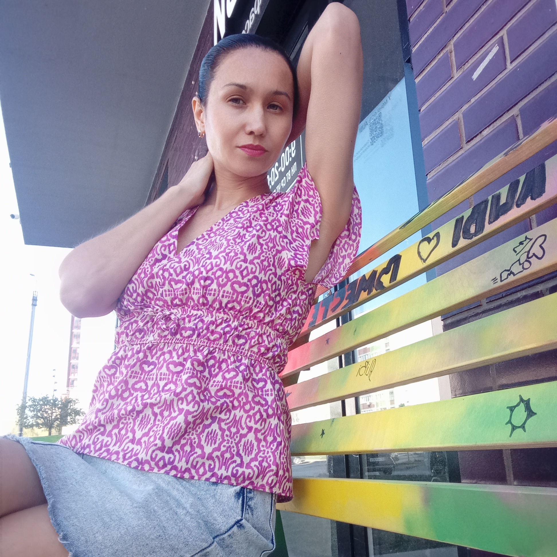 A woman is sitting on a brightly colored bench in Izhevsk, Russia. The bench has graffiti and drawings on it, including a heart and stylized letters. The woman is wearing a pink and white patterned top and a denim skirt. She has dark hair pulled back and is looking directly at the camera with one arm raised behind her head and the other hand resting on her neck. Behind the bench is a brick wall and a window with a sign displaying business hours. Buildings are visible in the background. The scene is lit by natural daylight.