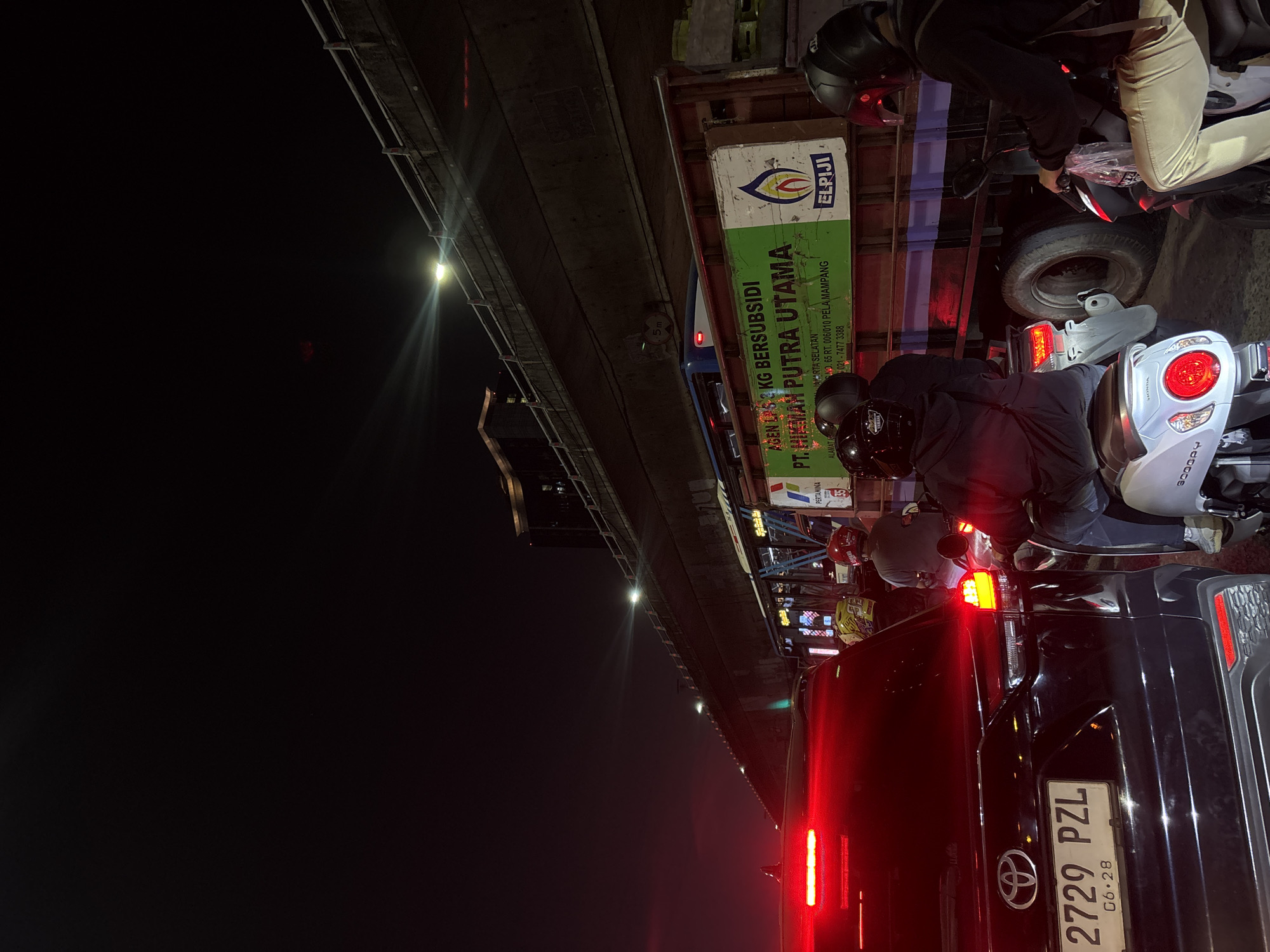 This is a night-time photograph taken in a traffic jam on an elevated road in West Jakarta, Indonesia. The scene is dominated by the glow of car and motorcycle lights against the dark sky. In the foreground, the rear of a black Toyota SUV is visible, displaying license plate "2729 PZL G6-28." To the right of the SUV, several individuals on motorcycles are present, their red taillights and helmets illuminated.
Behind the SUV, a large truck is stopped, featuring a green sign that reads "AGEN LPG & KG BERSUBSIDI PT. HIMAM PUTRA UTAMA." Below this, smaller text indicates "JAKARTA SELATAN," and an address with a phone number. The truck also displays the "ELPIJI" logo. A blue bus is partially visible behind the truck.
In the background, an elevated highway structure runs overhead, illuminated by streetlights. The overall impression is one of urban congestion at night, with vehicles and infrastructure typical of Jakarta.