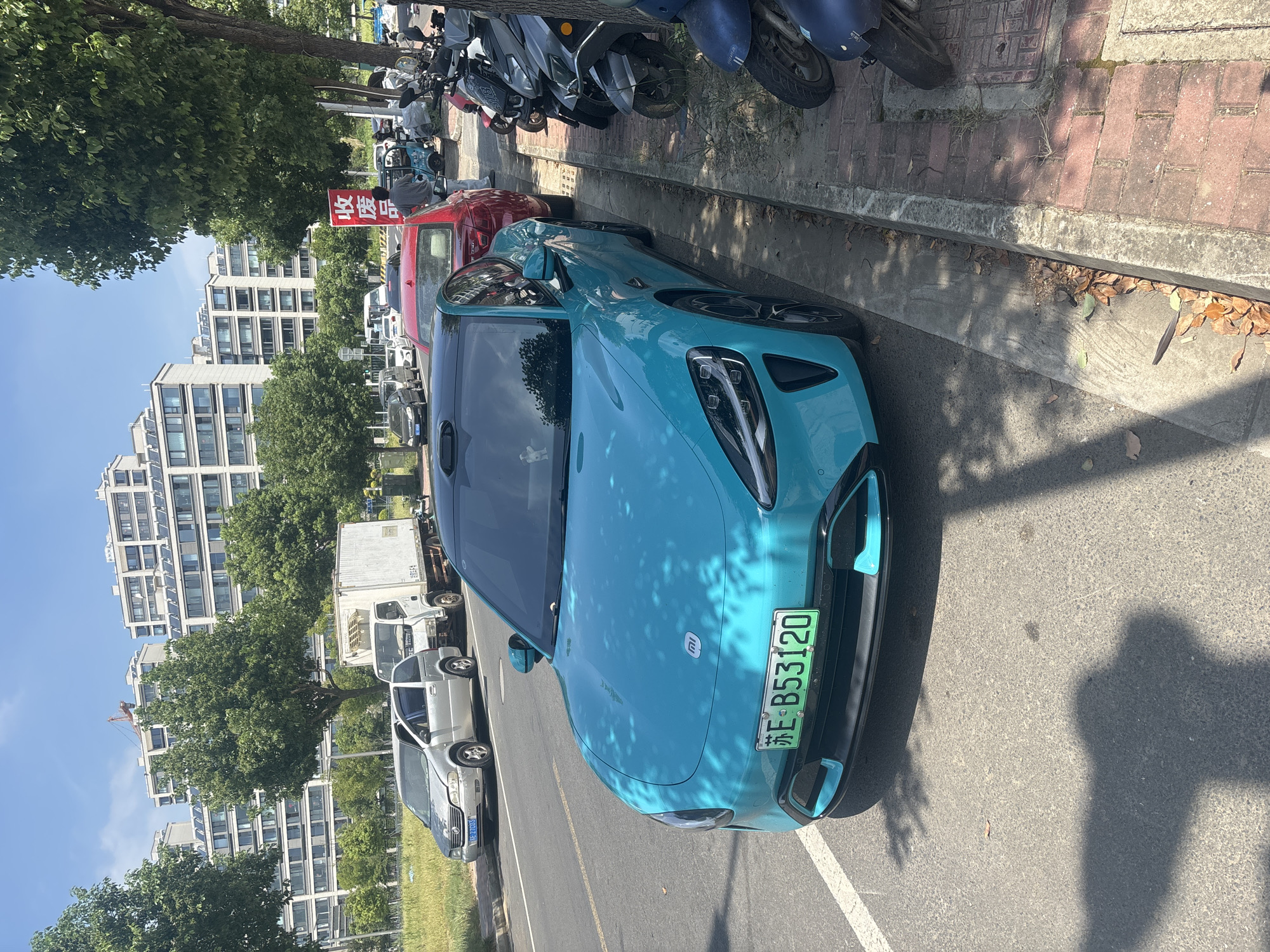 In Suzhou Industrial Park, China, a bright turquoise electric vehicle, likely a Xiaomi SU7, is parked on the right side of a street. The car's license plate reads "苏E B53120". To the left of the electric vehicle, a silver minivan and a white box truck are parked. The street is lined with trees and pedestrian sidewalks. Behind the vehicles, several multi-story residential buildings are visible under a clear blue sky. Several motorbikes and scooters are parked on the sidewalk to the right of the electric vehicle, and a red sign with Chinese characters is visible in the background. The scene depicts a typical urban streetscape in China, with a mix of vehicles, architecture, and infrastructure.