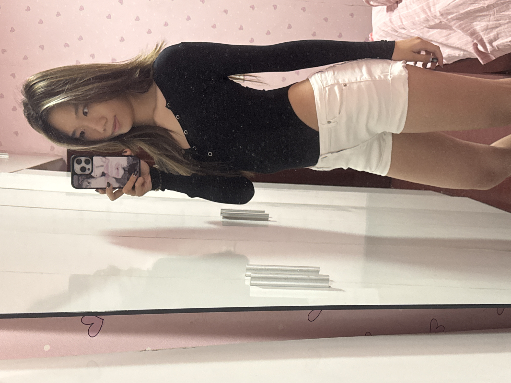 A woman is seen in a mirror selfie, wearing a black long-sleeved shirt and white shorts. She is holding a smartphone in her left hand. The background appears to be a room with pink wallpaper adorned with small hearts. A bed with pink and white striped bedding can be partially seen in the background. The location appears to be in a domestic interior, most likely a bedroom. There is no specific indicator of the exact city or region of Shapingba District, China within the image.
