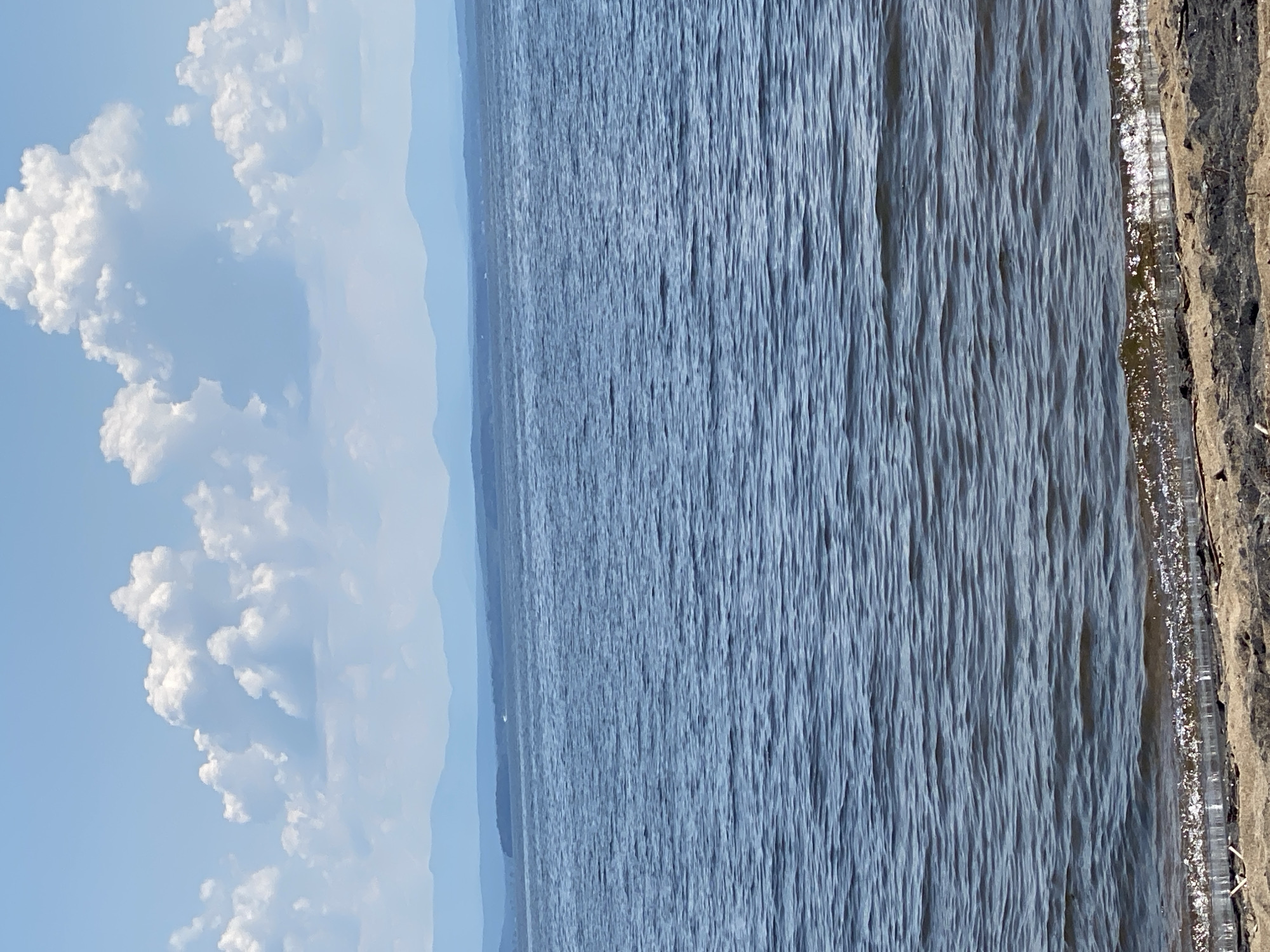 The image depicts a serene natural scene of a body of water, likely a lake or large river, with a distant shoreline and a bright sky with fluffy white clouds. The water is a deep blue and shows gentle ripples, suggesting a calm day. On the right side of the image, a textured embankment or shoreline is visible, with some dry vegetation or debris. The sky occupies the left portion of the image and is a light blue, adorned with large, puffy clouds that appear bright and illuminated, indicative of daylight. Faint, hazy mountains or landforms can be seen in the distance, blurring into the horizon. The perspective is from a slightly elevated position looking out over the water. There is no text visible in the image, and no people or specific objects are discernible. The overall mood is peaceful and tranquil, suggesting a pleasant day outdoors in a natural setting, possibly in the Town of Peru, USA. The time of day appears to be midday or afternoon, given the bright illumination and the appearance of the clouds.