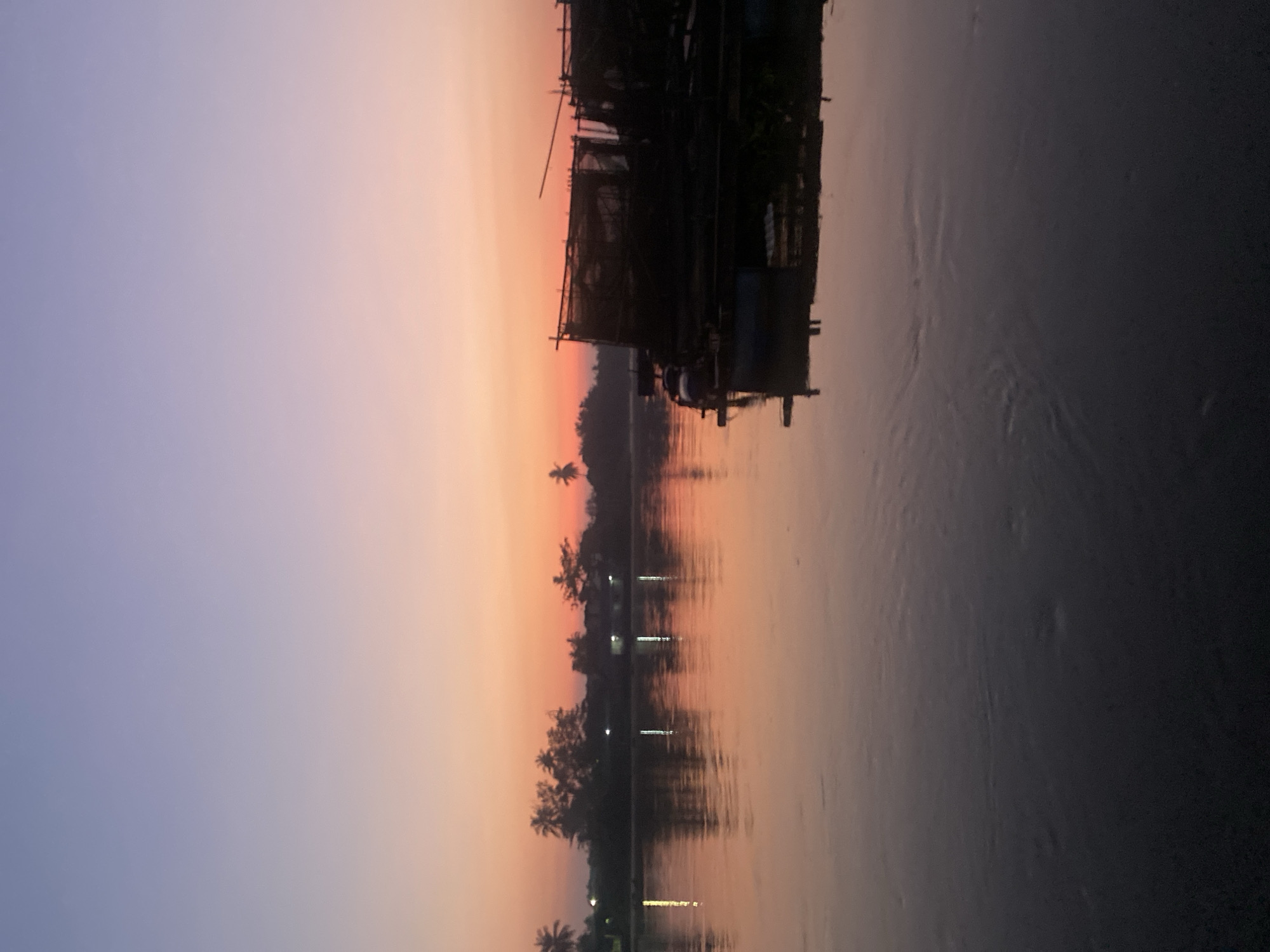 The image depicts a serene sunset over the Martapura River in Banjarmasin, Indonesia. The sky transitions from a soft lavender at the top to a vibrant orange and pink hue near the horizon, creating a beautiful gradient. Silhouetted against the colorful sky is a line of palm trees and vegetation along the riverbank, with a few faint lights suggesting buildings or homes.
In the foreground, the calm river reflects the colors of the sky and the objects on the water. On the right side of the river, several dark, silhouetted structures are visible, appearing to be houseboats or floating platforms with some form of netting or enclosures, possibly used for aquaculture or as dwellings. One structure has a person visible, tending to it or moving around. The water itself shows gentle ripples, distorting the reflections of the sky and the structures. The overall atmosphere is tranquil and peaceful, characteristic of a sunset scene in a riverside city.
