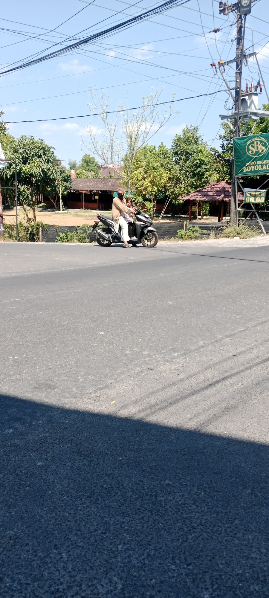 A man and a child are riding on a dark grey scooter on a paved road. The man is driving, wearing a tan shirt and light-colored pants. The child, wearing a white shirt and dark pants, is seated in front of him, holding onto his shoulders. The scooter is in motion, as indicated by the blurred background. The road is asphalt, with tire marks visible. In the background, there are trees with green foliage, some buildings with tiled roofs, and a utility pole with numerous wires. A green banner with white lettering is attached to the utility pole. The sky is clear and blue, with a few wispy clouds. The scene appears to be in a semi-urban or rural area.