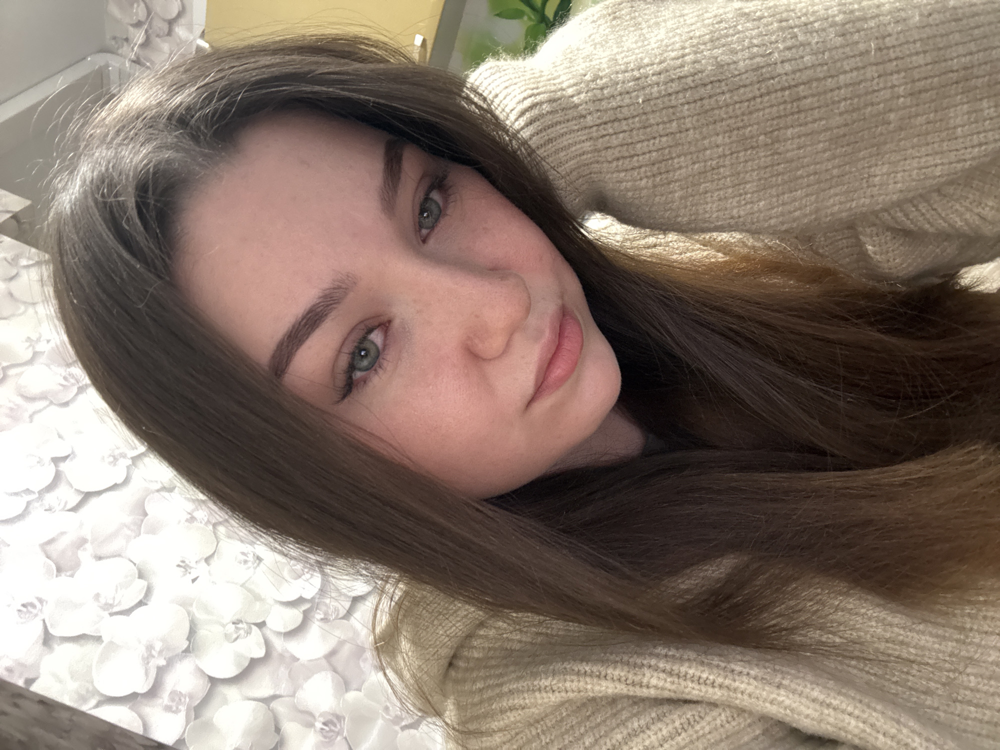 A young woman with long, dark brown hair and green eyes is shown in a close-up, head-and-shoulders shot. She is wearing a beige knitted sweater and leaning her head on her arm. The background is a wall with a floral wallpaper pattern in shades of white and grey. The lighting suggests an indoor setting. There is no discernible location in the city of Sakmary, Russia.