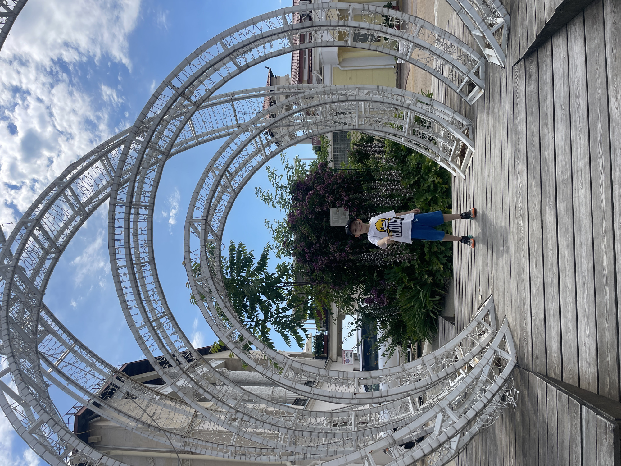 A young boy stands in the center of a large, white, metal arched structure on a wooden boardwalk. He is wearing a white t-shirt with a graphic of a yellow duck, blue shorts, and sneakers. Behind him, lush green bushes with purple flowers are visible, along with a building with light yellow walls and a red roof. The sky above is blue with scattered white clouds. The scene appears to be outdoors, possibly in a park or public space. This location is likely in Sochi, Russia, given the provided context.