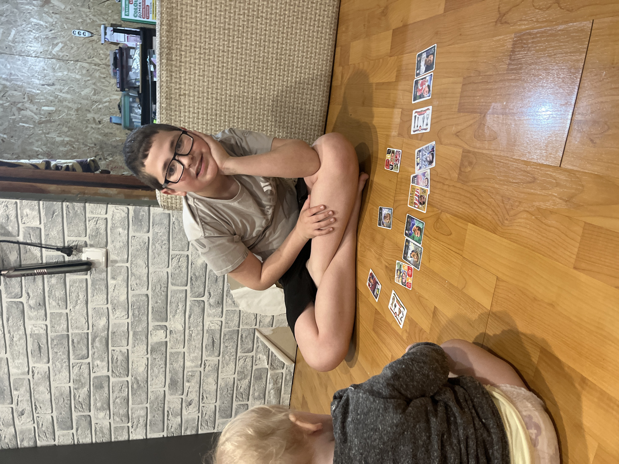 A medium-angle shot shows two children playing with soccer cards on a wooden floor, likely indoors. The older child, a boy wearing glasses and a gray t-shirt, is sitting cross-legged and facing the camera. He has his left hand resting on his face and a slight smile. The younger child, mostly obscured, is wearing a dark gray sweater. A line of soccer cards extends from the children towards the right side of the frame. The background consists of a gray brick-patterned wall on the left, a beige textured surface behind the older child, and a shelf unit with items on it in the top left corner. The location in the city of Vesyoloye, Russia is not evident in the image.