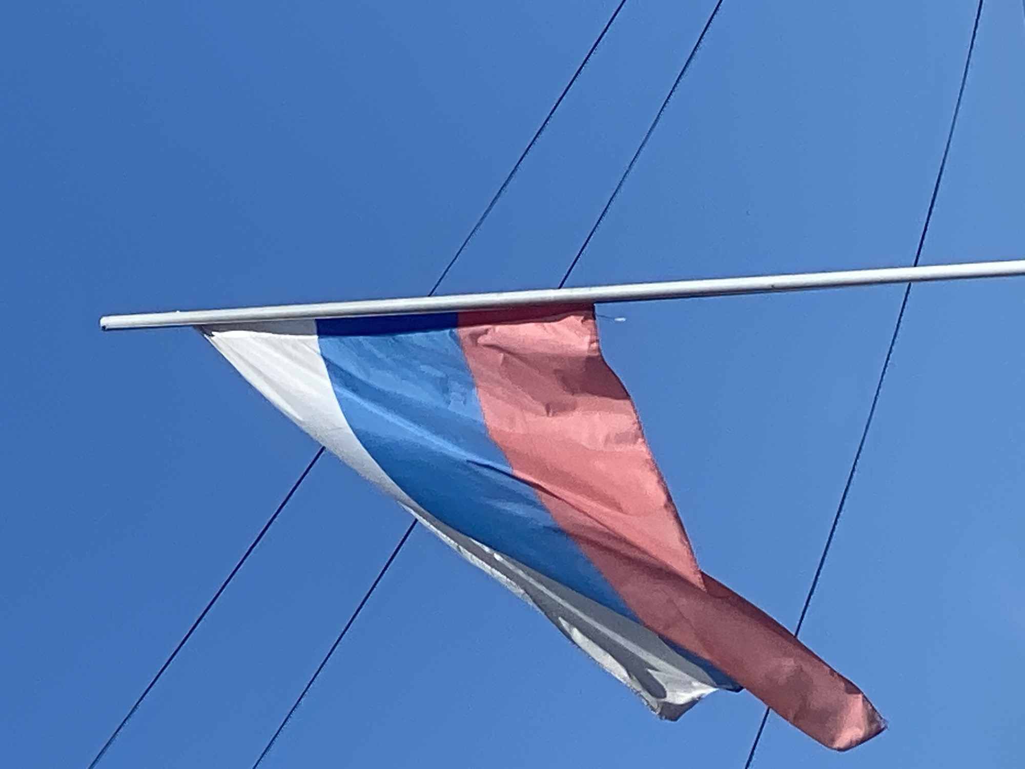 A Russian flag is fluttering in the wind against a clear blue sky. The flag is attached to a white pole on the right side of the image. Three dark wires are visible stretched horizontally across the sky in the background. The flag consists of three horizontal stripes: white on top, blue in the middle, and red on the bottom. The bottom of the flag is partially obscured by a wire. The image appears to be taken outdoors on a sunny day. The scene is devoid of people or specific landmarks that would indicate a precise location within Novokuznetsk.