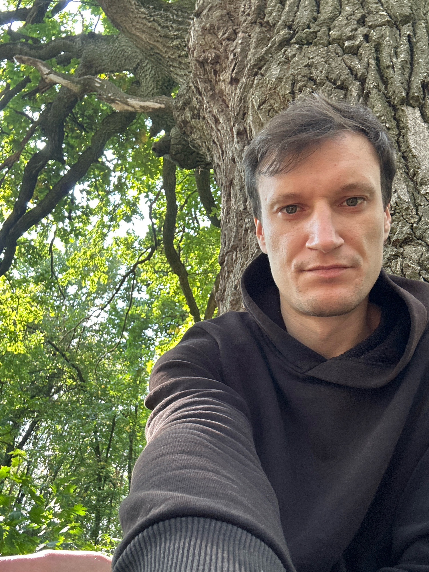 A man is taking a selfie outdoors, leaning against a large tree trunk. He is wearing a dark hooded sweatshirt. Behind him, the tree's branches extend upwards and are covered in green leaves, with sunlight filtering through. The setting appears to be a natural area with abundant foliage. There is no discernible urban landscape or specific landmarks to indicate a location within Makiv, Ukraine.