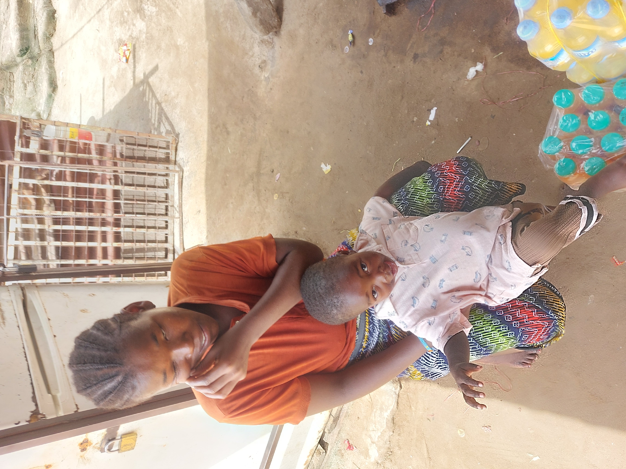 A woman in an orange t-shirt and a patterned skirt is holding a child in a light pink shirt and brown pants. The woman is looking at the camera and smiling, while the child is looking away. They are sitting on a tan concrete surface next to a stack of water bottles. A metal grate is visible on the left side of the image. The location appears to be outdoors in an urban setting, but further specifics within Dar es-Salaam are not discernible.