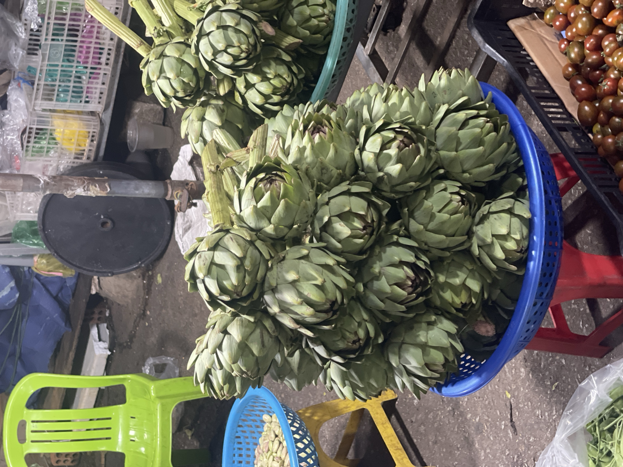 The image shows a street-level view of a market scene. Several baskets contain artichokes of varying sizes, with a few tomatoes visible in another basket, presumably for sale. The baskets are of different colors, including blue and green. Additional elements in the environment include a green plastic chair, another blue basket, and what appears to be a metal bar. The lack of people suggests a pause in market activity or a focus on the products themselves. Considering the focus on artichokes, it's plausible this photo was taken in or around Da Lat, Vietnam, an area known for its artichoke cultivation.