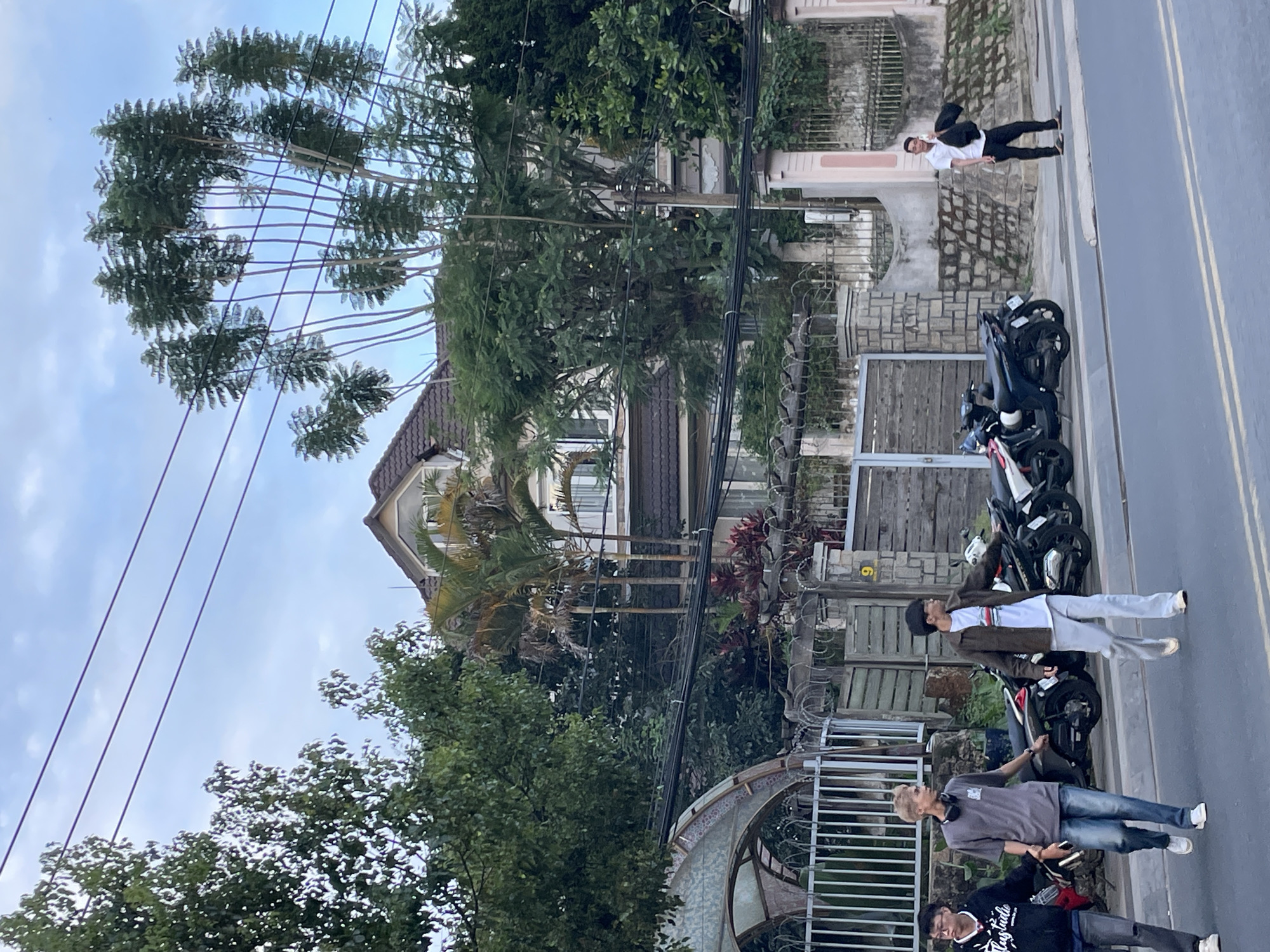 In the city of Da Lat, Vietnam, a street scene unfolds with a large, multi-story house as the backdrop. The house features a prominent, steeply pitched roof with dormer windows and is surrounded by lush greenery, including tall trees and palm trees. A grey concrete fence with decorative metalwork runs along the property line, topped with barbed wire. Adjacent to the fence, wooden gates are visible.
On the street in front of the house, four young men are present. Two men, one wearing a black hoodie and the other a grey t-shirt and jeans, walk side-by-side. Another man in a brown jacket and light-colored pants walks further down the street, looking to his right. Further down the sidewalk, a fourth man in a white polo shirt and black pants stands holding a black bag. Several motorcycles are parked along the roadside. The sky above is partly cloudy, with visible power lines running across the frame.