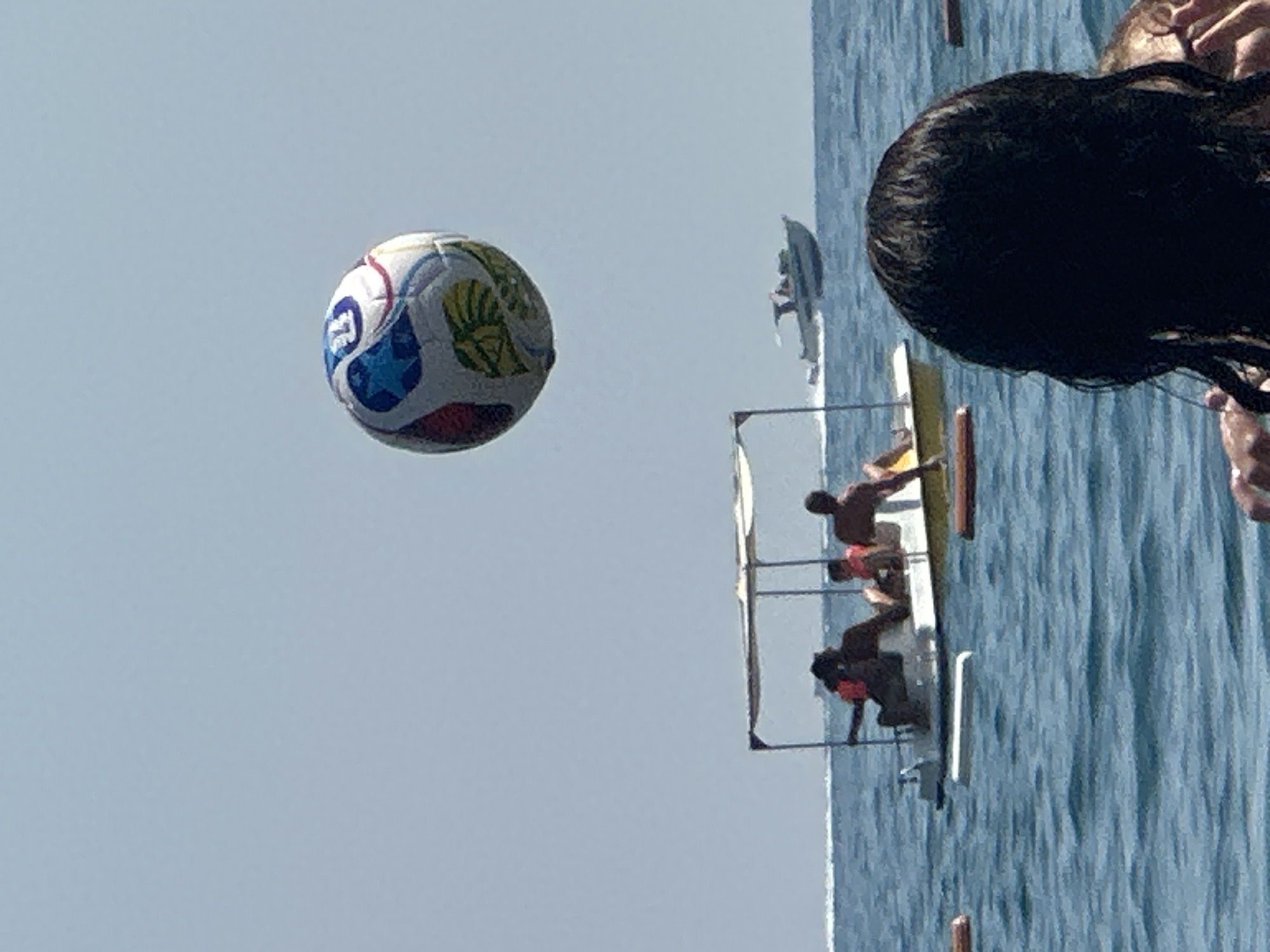 An overhead view of a beach scene in Boreti, Montenegro, features a large, colorful soccer ball suspended in the sky. In the middle ground, a small, flat-bottomed boat with a canopy is occupied by several individuals wearing life vests. A white speedboat is visible in the distance, moving across the blue water. The foreground is partially obscured by the back of a person's head, their dark hair damp. The ocean is calm and the sky is clear.