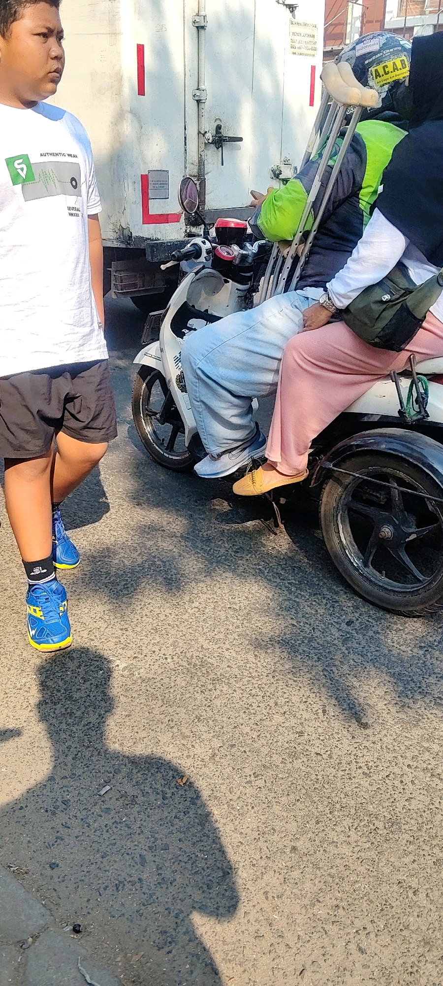 The image depicts a street scene in Cibatu, Indonesia, featuring several individuals and a motorcycle. In the foreground, a person wearing a white t-shirt and black shorts stands on the asphalt road, casting a shadow. They are wearing blue and yellow athletic shoes with black socks. Behind this person, a motorcycle is parked. On the motorcycle, two individuals are seated. The person in the front, likely the driver, is wearing light blue jeans and grey sneakers. They are holding onto the handlebars and appear to be wearing a helmet with stickers, including one that says "Indonesia" and "A.C.A.B." They are also carrying crutches. The person seated behind them is wearing a white long-sleeved shirt and pink pants, with a dark green bag slung across their body. The background shows the side of a white truck or shipping container, with some red markings and a latch. The ground is paved with asphalt and appears to be a public road. The lighting suggests it is daytime, with shadows cast by the people and the motorcycle.