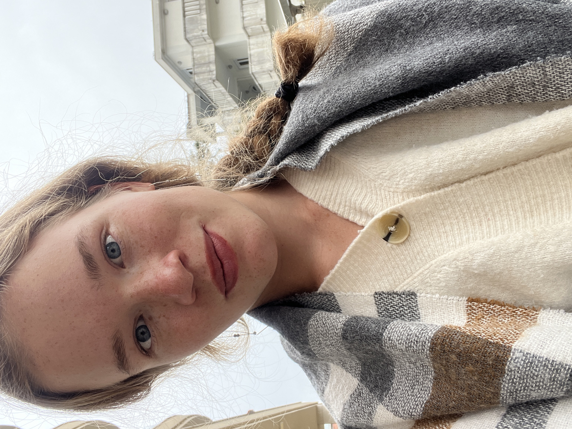A selfie, taken at an upward angle, shows a fair-skinned woman with freckles and blue eyes wearing a cream-colored cardigan with a single visible button and a gray, white, and brown plaid shawl. Her hair is blonde near the front and brown at the back, pulled into a ponytail with a black band. Behind her, a modern, multi-story building is visible against a light sky. The setting appears to be Budva, Montenegro, given the distinctive architectural style of the building in the background. A partial view of a low, light-colored barrier can be seen in the bottom corner of the frame.