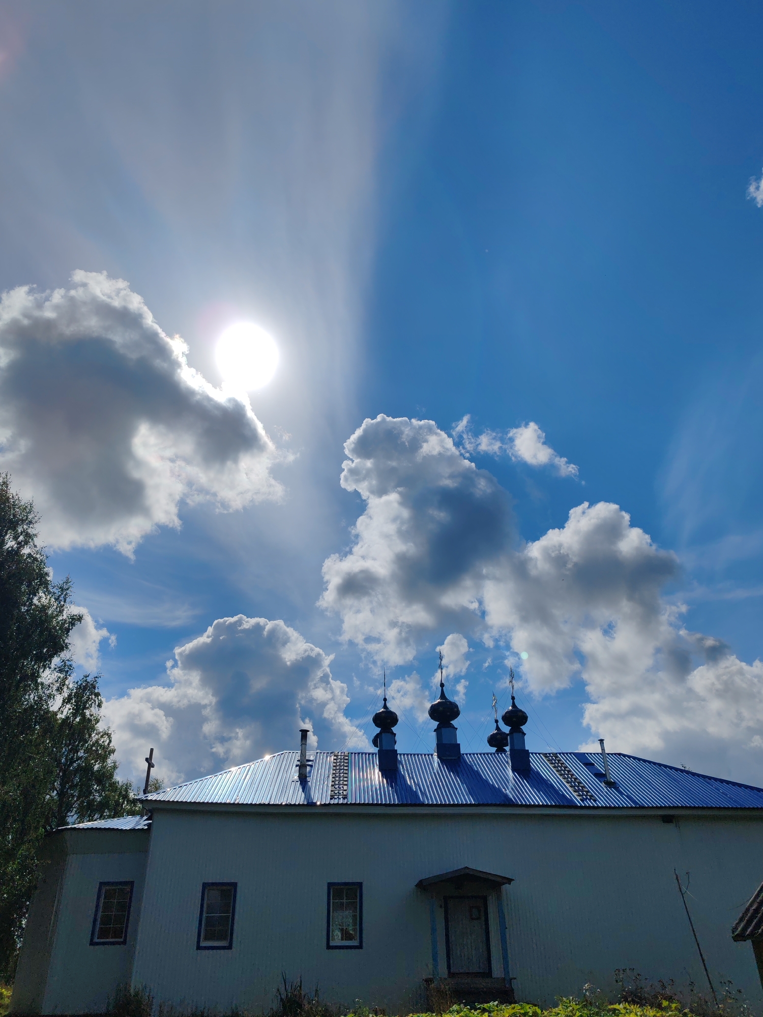 The image captures a low-angle view of a white, rectangular building with a blue, corrugated metal roof, likely a church or religious structure, in the city of Chukhlem, Russia. Four dark, onion-shaped domes with finials are visible on the roof, along with a chimney. A small, covered entrance with a door is situated on the front of the building, flanked by three rectangular windows with dark blue trim. A cross is visible atop a portion of the roof to the left of the domes. To the left of the building, the upper branches of a dense green tree are seen. The sky above is a vibrant blue, with a bright sun partially obscured by white and grey cumulus clouds. Sunlight beams emanate from the sun, creating a bright, atmospheric effect. The foreground shows some dry grass and vegetation.