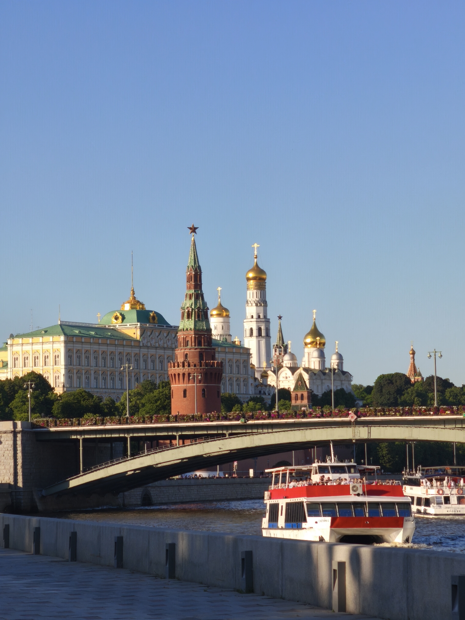 The image displays a clear, sunny daytime scene of the Moskva River and the Moscow Kremlin complex in Moscow, Russia.
In the foreground, a smooth, light grey concrete embankment runs along the lower left of the frame. The river surface shows ripples and a dark reflection under the midground bridge. A white and red passenger boat, with "СНЕЖАНА" (Snezana) visible on its side, moves from the left towards the right, creating a distinct wake. Several passengers are visible on its upper deck. Further back and to the right on the river, a second, smaller white passenger boat, with "КРОКУС" (Krokus) partially visible, also moves rightward.
In the midground, a multi-arched bridge, likely the Bolshoy Kamenny Bridge, spans the river. Its support structures are made of light grey stone, and the main arch is a green metal truss. The bridge deck is adorned with numerous flower boxes filled with red and purple blossoms. Trees and dense green foliage line the riverbank behind the bridge.
The background is dominated by the iconic Moscow Kremlin. On the left, a large, ornate yellow palace building with a green roof and golden domes is prominent. To its right, a tall, red brick tower with a green conical spire, topped by a prominent red five-pointed star, stands out. Further right, a complex of white buildings features multiple golden-domed cathedrals and a tall, white bell tower with a golden dome and cross. Beyond these structures, another smaller, red-roofed building with an intricate design is visible. The sky above is a uniform, clear light blue with no clouds. The lighting suggests a bright, possibly late afternoon or early morning sun, casting long shadows on the embankment and illuminating the Kremlin structures warmly.