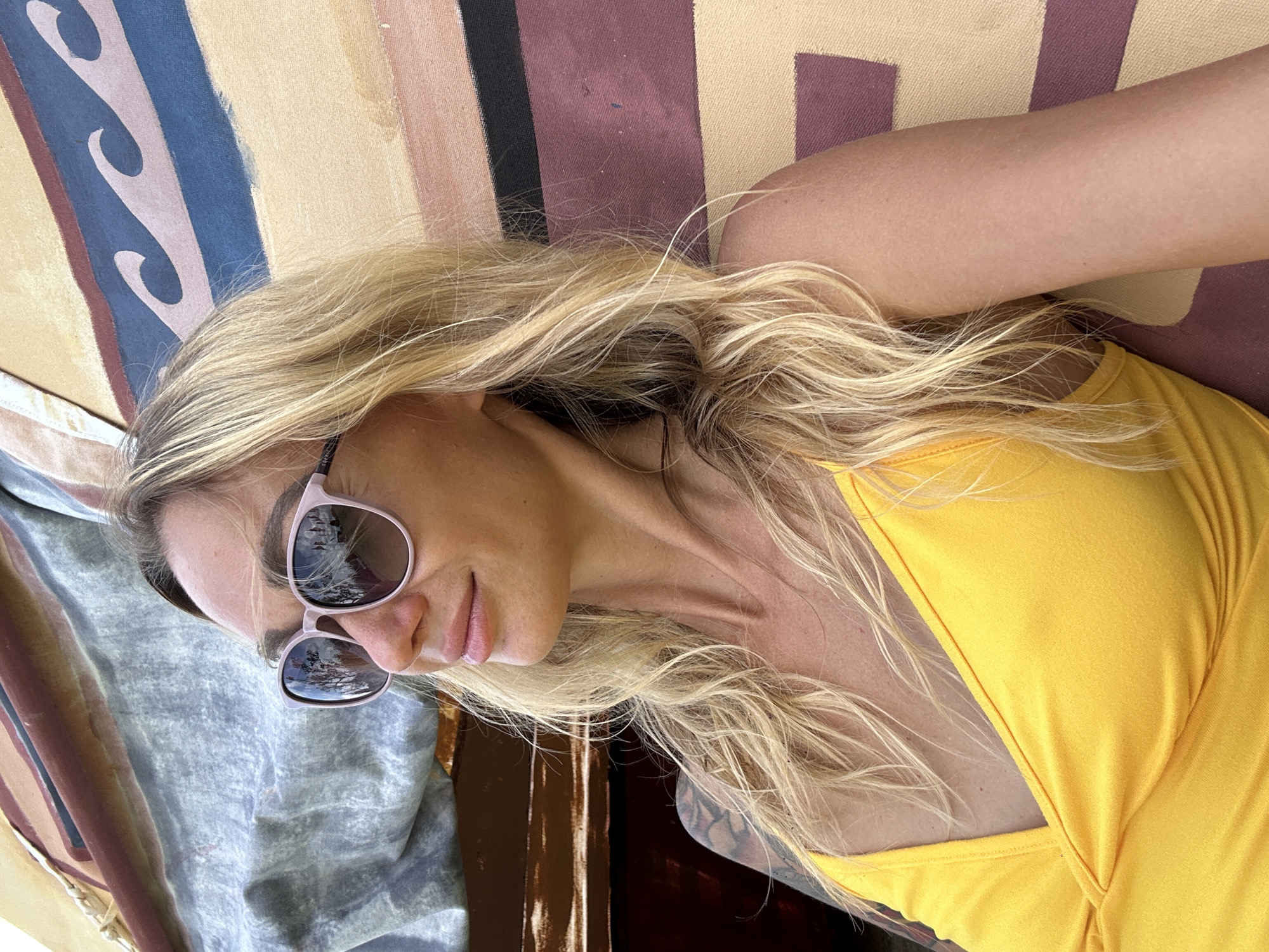 This media file features a close-up, vertically oriented, head-and-shoulders shot of a blonde woman wearing sunglasses and a yellow top. She is lying down, with her head resting on a surface that appears to be fabric with a striped and abstract pattern. Her sunglasses reflect trees and what looks like a cloudy sky. Her hair is blonde and windswept, falling around her face and shoulders. The woman has a relaxed and possibly content expression. The lighting suggests it is daytime, likely outdoors, given the reflections in her sunglasses. The background behind her is a muted, abstract pattern of stripes in various earthy tones and blue. There is no visible text in the image. The location context provided is Thôn 3, Vietnam, but this is not directly inferable from the visual content itself.