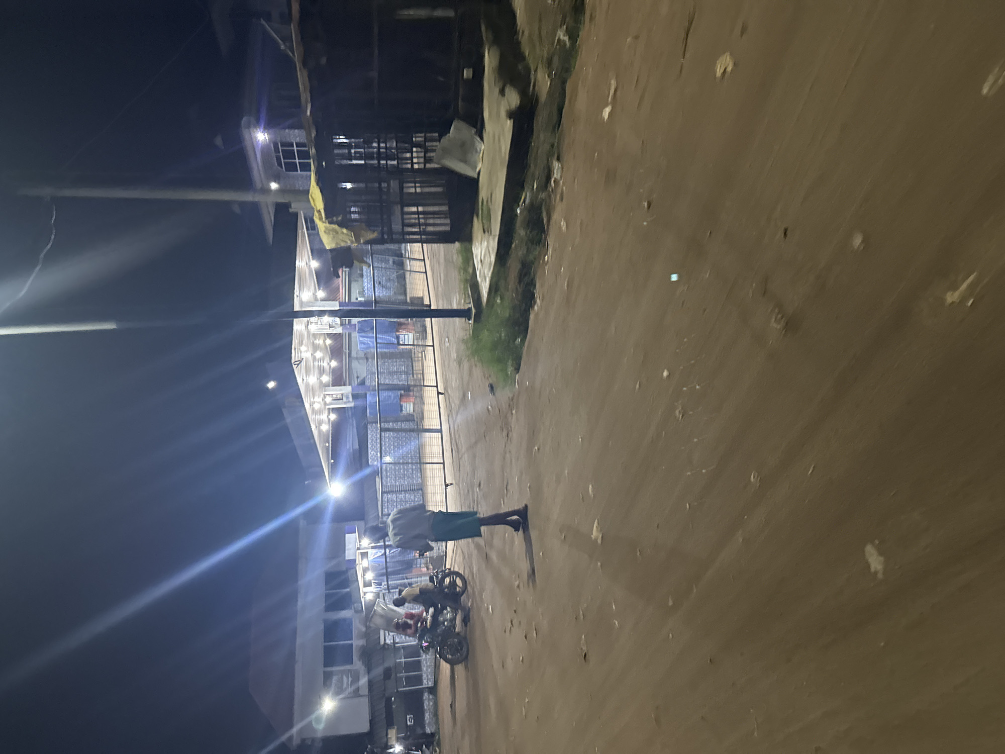 At night in Ihiagwa, Nigeria, a street scene shows a man standing near two motorcycles. The scene is lit by streetlights. One building is lit up with a light-strung patio.