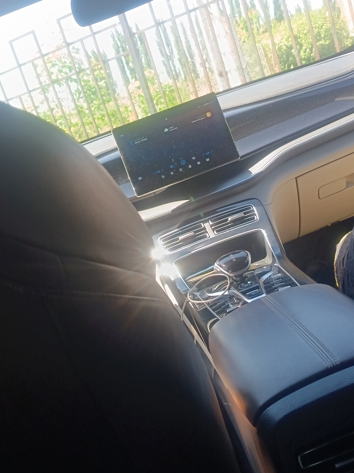 The image displays the interior of a vehicle, captured from a rear passenger's vantage point, looking forward towards the front cabin. The prominent foreground on the left is occupied by the dark, upholstered back of a front passenger seat. Intense sunlight streams into the vehicle, generating a lens flare effect above the center console.
The dashboard features a large, rectangular digital infotainment screen displaying a user interface with various icons, text, and a digital clock set to "10:44". The screen's surface is noticeably dusty. Below the screen are horizontal air vents accented with silver-colored trim. The center console includes a black, rounded electronic gear selector, marked with "P" for Park. To the left of the selector, an open compartment contains a pair of eyeglasses and a white charging cable. Below the gear selector, a control panel with dials and buttons, also with silver trim, is visible. A dark, stitched leather or synthetic leather armrest is positioned on the right side of the center console. Further right, a light-colored glove compartment is present, and a foot, partially clad in dark material, is visible in the passenger footwell.
Through the front windshield, a bright outdoor environment is discernible. A light-colored metal fence, characterized by a grid pattern, is prominently featured. Behind the fence, green foliage from trees and bushes is observed beneath a clear, bright sky, indicating daytime. No individuals are fully visible, though the partial view of a foot suggests a front passenger is present. No explicit actions are depicted. The location is Unknown, Unknown.