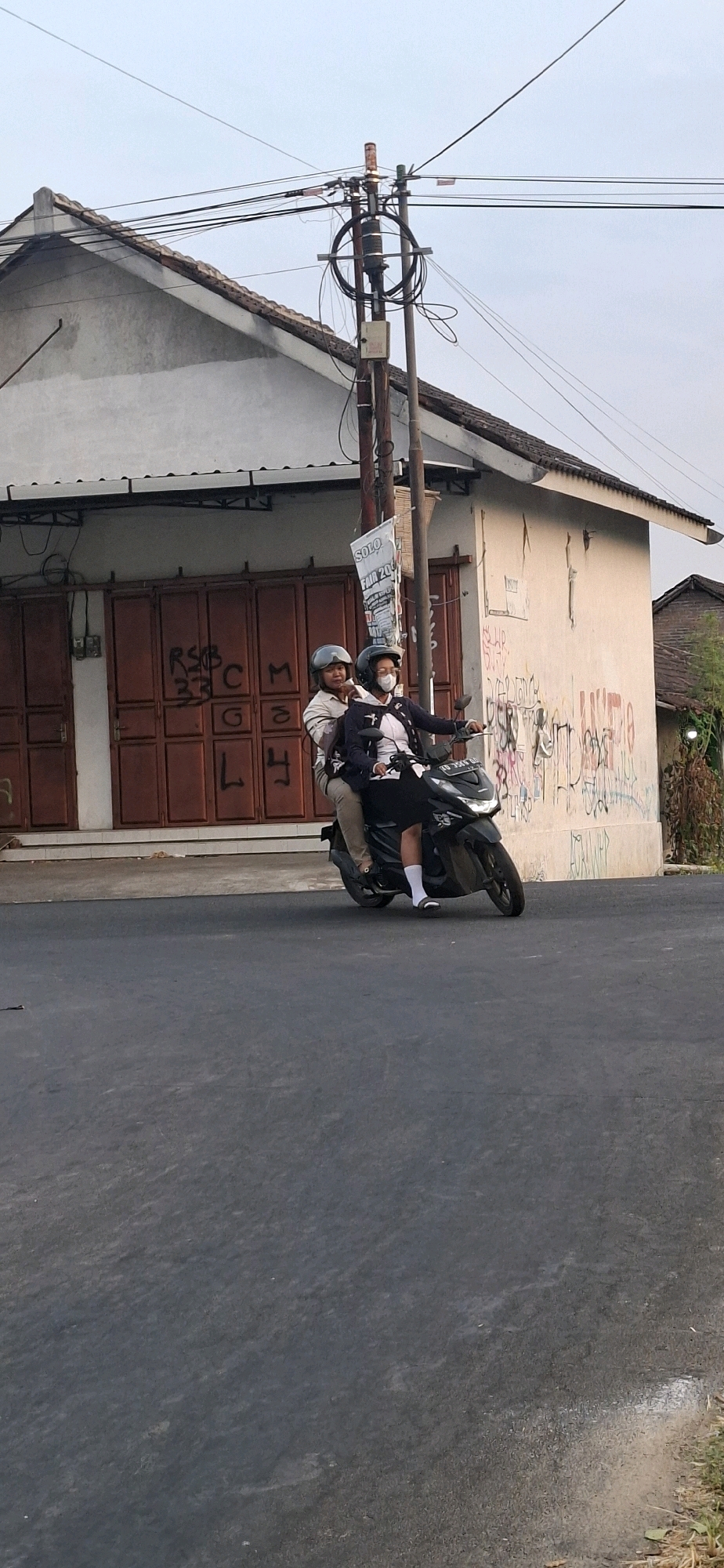 In Desa Dibal, Indonesia, two individuals are riding a black scooter on a paved road. The person in front, wearing a light-colored shirt and helmet, is the driver. The person behind, wearing a dark jacket and mask, appears to be a passenger. They are traveling past a building with a white facade marked with graffiti and large brown double doors. A utility pole with numerous wires stands to the right of the building, and a poster advertising "SOLO FAIR 2015" is attached to it. The sky is overcast, and the overall lighting suggests it is either early morning or late afternoon. The road surface is asphalt, and there is some dry vegetation visible on the right side of the frame.
