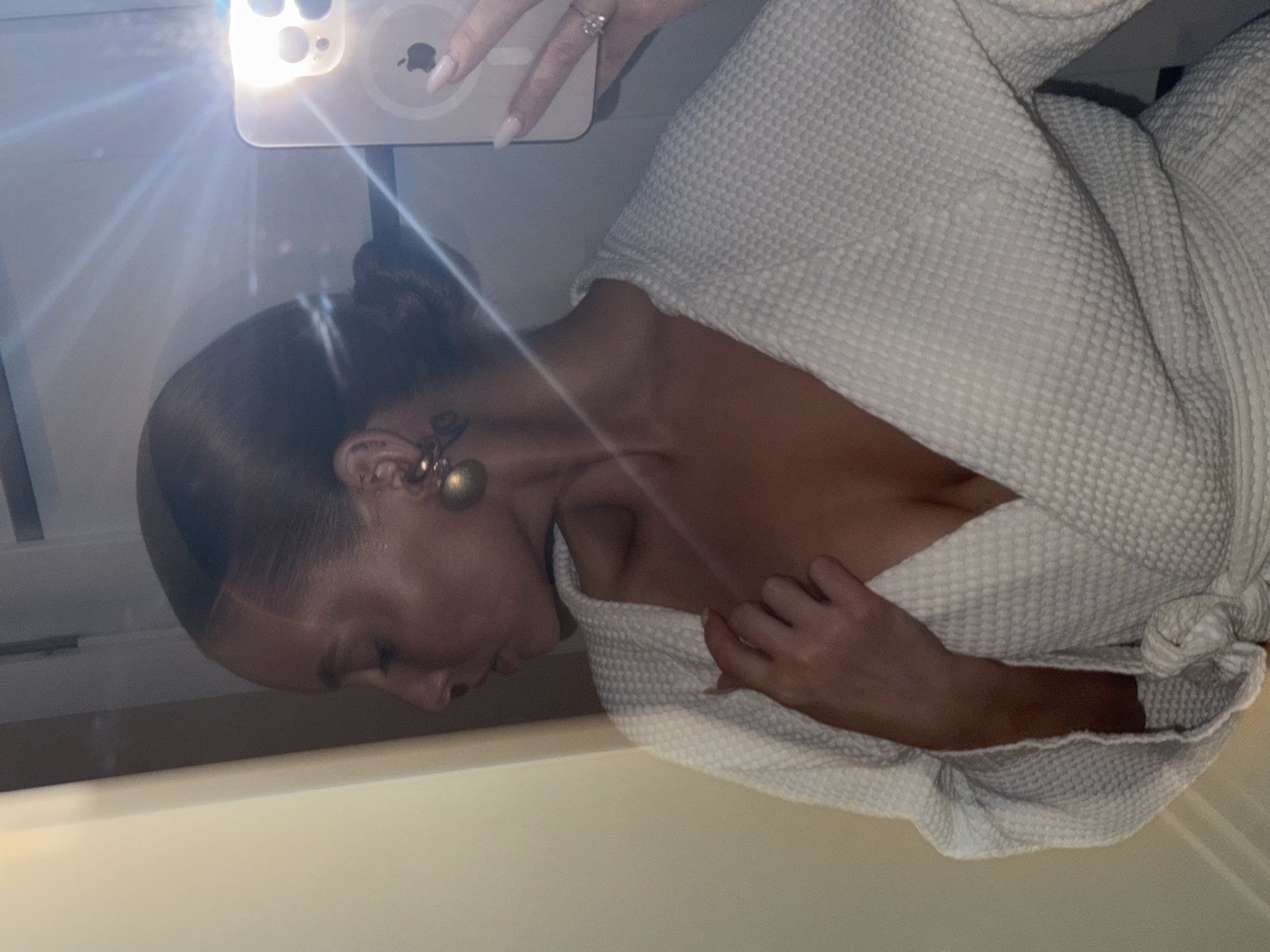 A woman in a white waffle-weave robe is taking a selfie. She is holding a smartphone up to the mirror. The selfie flash is on. She has light brown hair pulled back in a bun, and is wearing gold earrings. She has a tattoo behind her ear. The background is blurred, but appears to be a light-colored wall. There are no discernible indications that place the image in Chaikovsky, Russia.