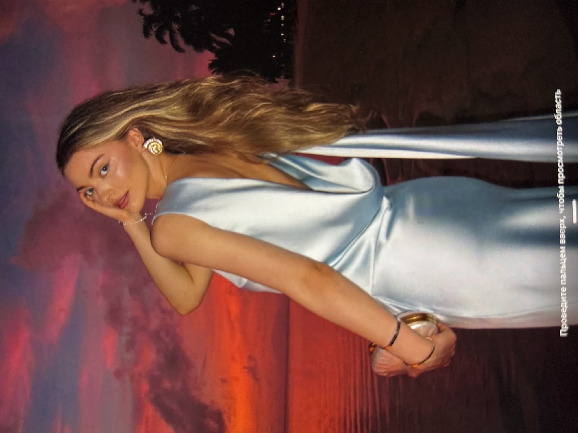 A woman is pictured in a light blue satin gown, with her back to the camera, holding a clutch purse. Her left arm is raised, with her hand touching the back of her head. She is wearing a bracelet and a large, ornate earring on her right ear. The background is a vibrant sunset over a body of water, with palm trees silhouetted against the sky on the right. The scene is outdoors, possibly at a beach resort. The location is not Găvănoasa, Moldova.