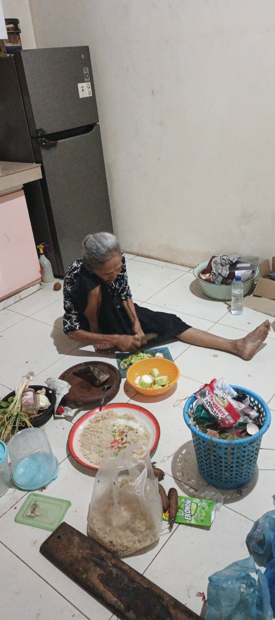 In Kasiyan, Indonesia, an elderly woman with grey hair is sitting on a tiled floor, preparing food. She is wearing a black and white patterned top and a black skirt. In front of her, she is cutting vegetables with a knife on a blue cutting board. Various food items and kitchen utensils are scattered around her, including a wooden chopping board, bowls of chopped vegetables and what appears to be rice or grain, a plastic bag filled with a similar granular food, a blue plastic basket containing various wrappers and food scraps, and a bottle of water. The scene is indoors, with a large grey refrigerator visible to the left and a plain wall behind her. The overall environment suggests a domestic setting, possibly a kitchen area.