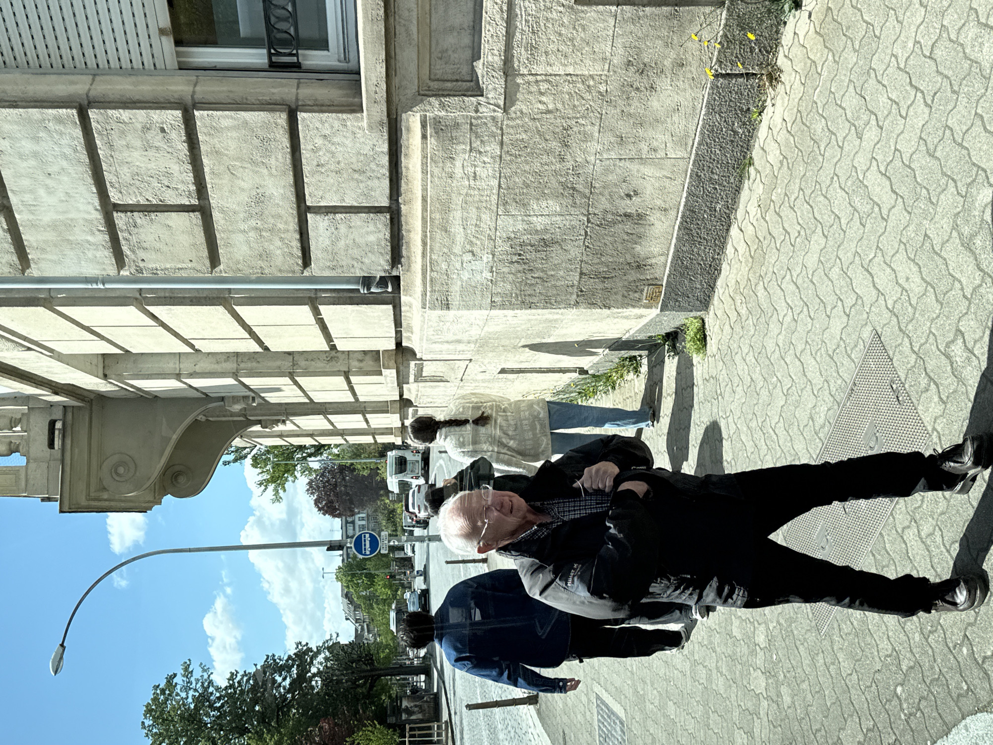 This image depicts a sunny urban street scene in Strasbourg, France. In the foreground, an older man with white hair and glasses, dressed in a dark jacket and pants, walks on a textured grey sidewalk with interlocking pavers. He is looking towards the viewer's right, and strong sunlight creates clear shadows on the ground.
Behind the prominent figure, other pedestrians are visible: one person in a blue jacket partially obscured, and another in a light-colored coat further back. The background reveals a street with several cars and a white bus, suggesting an active urban environment.
To the left, a multi-story building with a light-colored stone facade features ornate architectural details, including decorative corbels and windows with shutters. A grey pipe runs along the building's exterior. Above the street, a curved street light extends into a bright blue sky dotted with scattered white clouds. A blue circular road sign with a white bus icon is visible in the mid-ground, indicating a dedicated bus lane or stop. Small patches of greenery and trees line the street, contributing to the city landscape. The overall atmosphere is bright and bustling under clear, sunny weather.