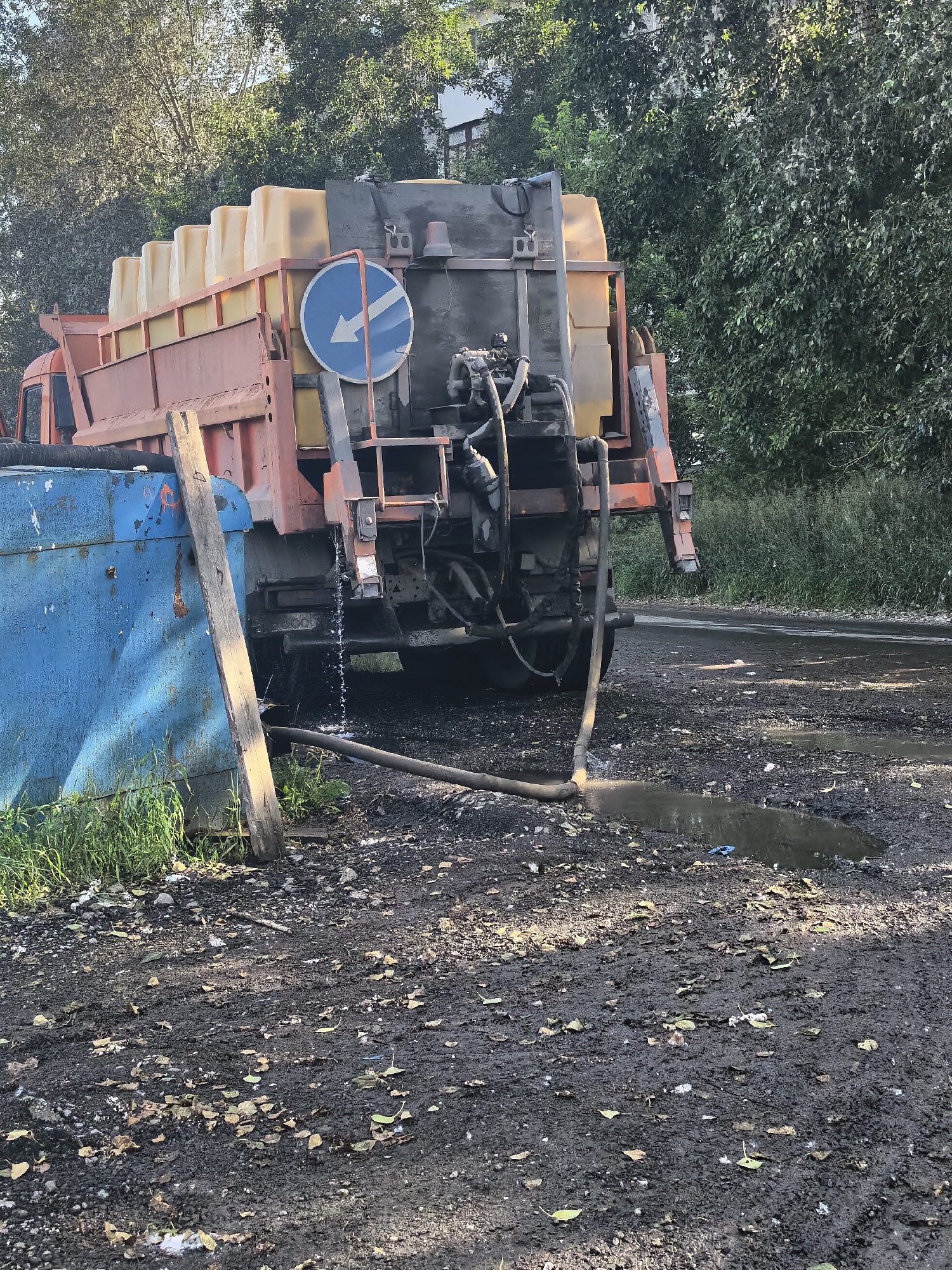 The image shows a rear view of a large orange truck with a grey tank filled with yellow containers. A blue sign with a white arrow pointing left is attached to the side of the truck. A hose is connected to the back of the truck and extends onto the ground, with a small amount of liquid appearing to be leaking from it and forming a puddle. Next to the truck is a large blue dumpster. The ground is covered in dark dirt and some scattered leaves. Trees and foliage are visible in the background, and a building can be seen through the trees. The scene appears to be outdoors, possibly in a work or industrial area, given the presence of the truck and dumpster. The lighting suggests it is daytime, with sunlight filtering through the trees. There is no visible text in the image.