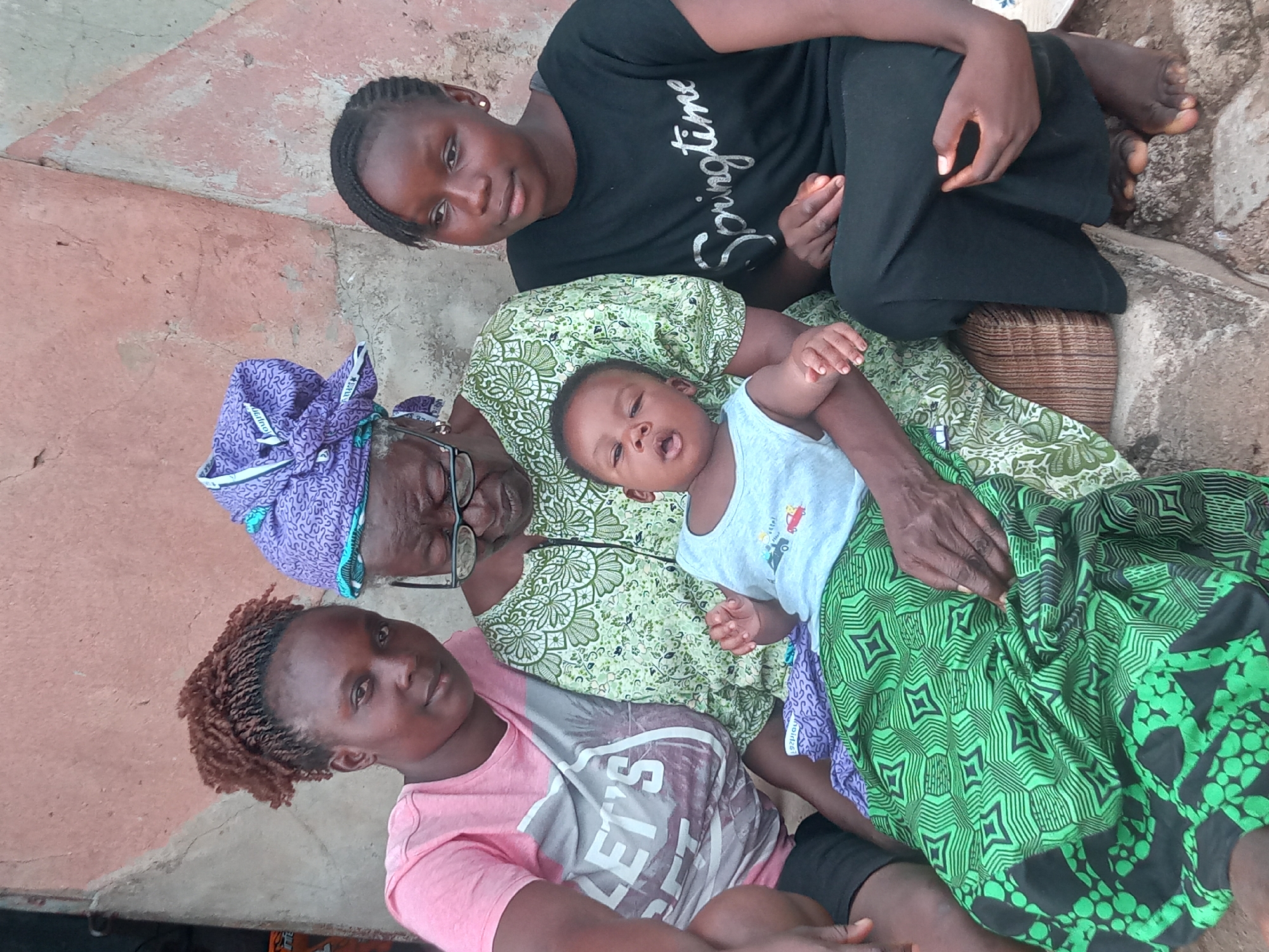 In Sabon Tasha, Nigeria, an elderly woman with glasses and a purple head wrap sits on the ground with a baby lying on her lap. The baby wears a light blue shirt. A young woman in a pink "LET'S" shirt sits beside the elderly woman. Another young woman in a black "Springtime" shirt sits above them. They are all on a woven mat on a concrete surface.