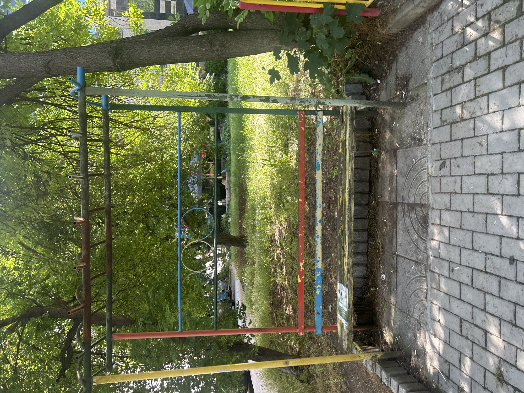 The image depicts an outdoor scene in Varna, Bulgaria, featuring a worn, colorful playground structure. The structure is primarily made of metal poles painted in yellow, red, blue, and green. A wooden bench, weathered and chipped, is attached to the structure. Above the bench, horizontal bars are arranged, with some ending in pipe-like fittings. In the background, there is abundant greenery, including trees with lush foliage and overgrown grass, suggesting a slightly unkempt urban park or courtyard area. Some distant vehicles and buildings are visible through the trees. The ground in the foreground is paved with grey bricks and stone tiles. Cigarette butts are scattered on the ground around the structure and on the paved areas. The lighting suggests it is daytime with dappled sunlight filtering through the leaves.
