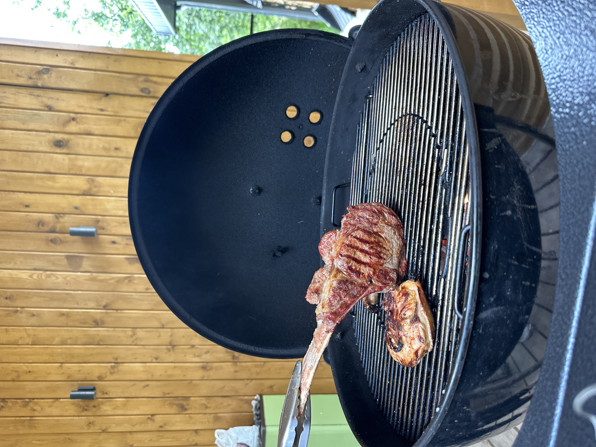 A tomahawk steak and a smaller cut of meat are being grilled on a black Weber charcoal grill. The grill is open, and the steaks are positioned on the grates over glowing charcoal. A pair of metal tongs is holding the bone of the tomahawk steak, as if to turn it. The grill is situated on a wooden deck with a slatted railing. In the background, a light-colored wooden wall with two modern sconces is visible. Further back, some greenery and a portion of a building's roof suggest an outdoor setting. The location is the city of Perm, Russia.
