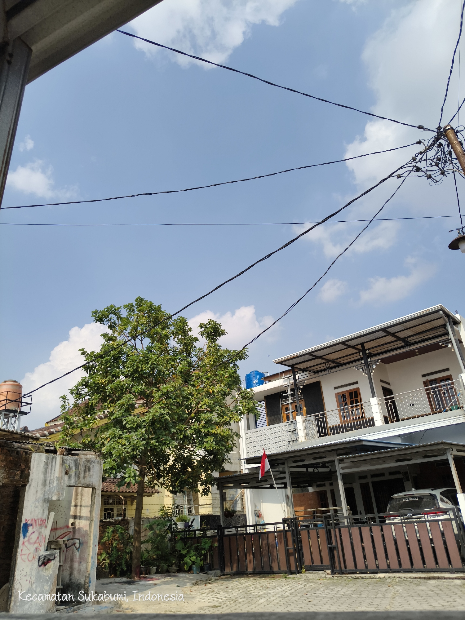 The image captures a street scene in Sukabumi District, Indonesia, under a bright blue sky with scattered white clouds. Overhead, a complex network of black electrical wires crisscrosses the frame, supported by a utility pole on the right. In the foreground, a paved road leads towards a modern two-story white house with a metal-roofed carport. The house features wooden-framed windows and doors, and a balcony on the upper floor. A white car is parked under the carport. To the left of the house, a lush green tree stands tall. Adjacent to the tree, a partially visible weathered concrete structure with graffiti is present. Further to the left, a water tank is visible on a metal frame atop a building. The scene is static, with no people or animals actively engaged in any discernible actions. The environment suggests a residential area in an urban or semi-urban setting.