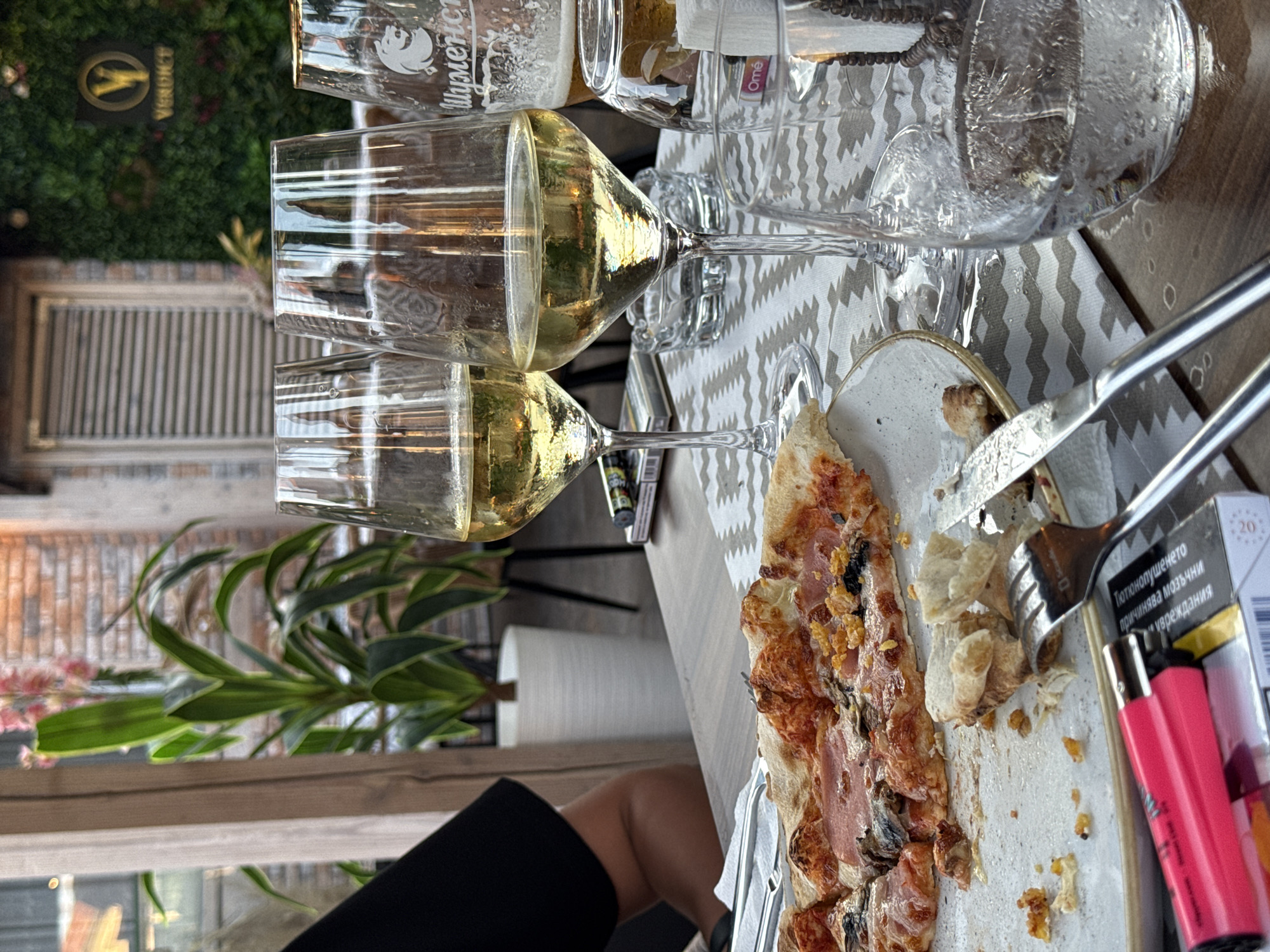 The image shows a table with drinks and food, likely at an outdoor dining establishment in Ravda, Bulgaria. Two wine glasses filled with a golden liquid are prominently displayed, suggesting a meal or a break. In the background, a lush green wall, possibly artificial foliage, and a wooden door or window add to the setting's ambiance. A partially eaten pizza lies on a plate, with a fork and knife nearby, indicating that a meal is in progress or has recently concluded.
Several other objects are scattered on the table. A glass bottle filled with water and ice sits next to the wine glasses. Two brightly colored lighters, one pink and one red, are placed near a pack of cigarettes, hinting at the presence of smokers. A printed brochure or packaging with Cyrillic text is also visible. The overall scene appears casual and relaxed.
The lighting suggests it might be daytime, with natural light illuminating the scene. The presence of plants and the outdoor setting imply a pleasant weather day. The focus on food and drinks, combined with the casual arrangement of items, evokes a sense of leisure and enjoyment.