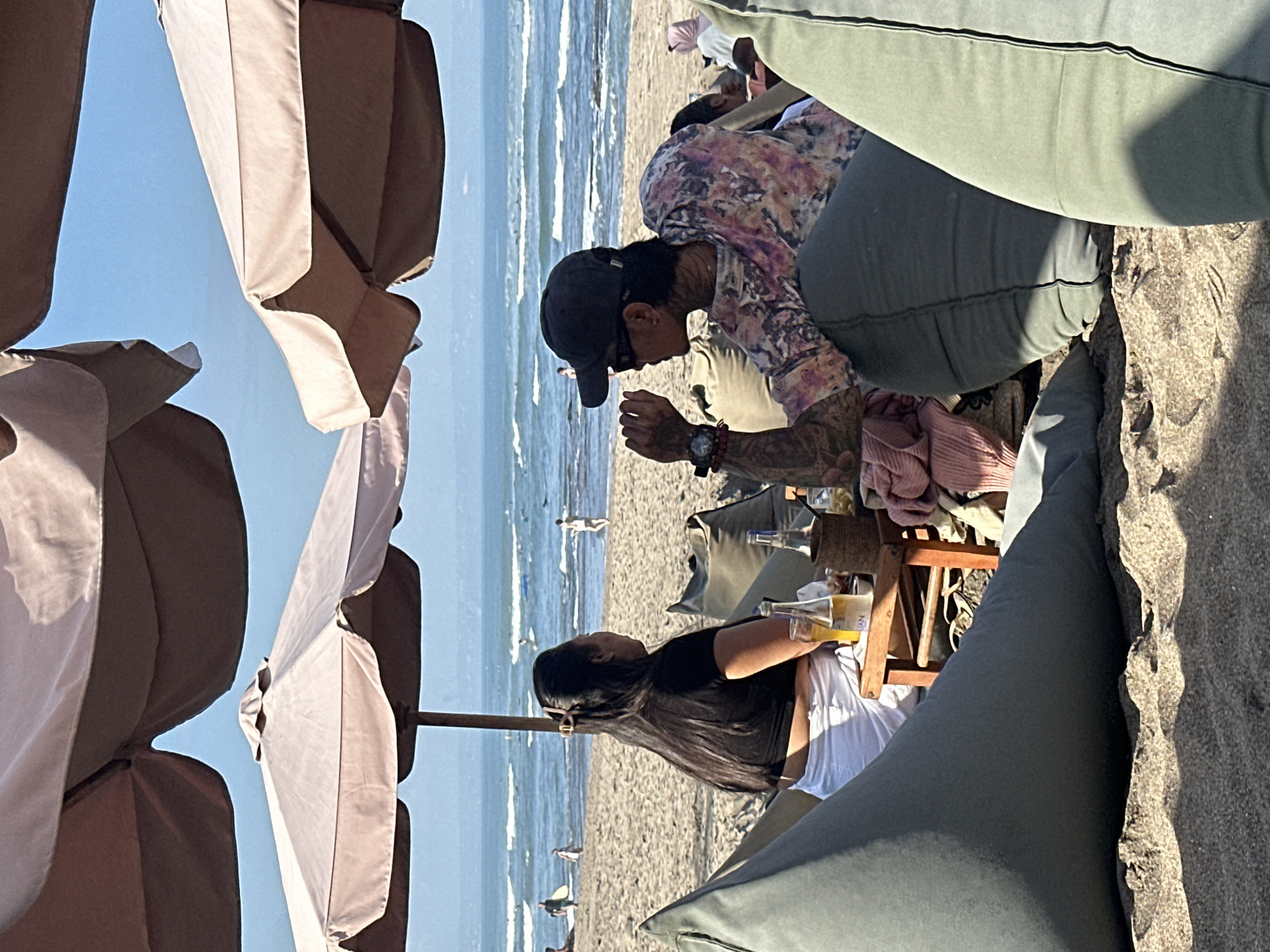 The image depicts a serene beach scene in North Kuta, Indonesia, under a clear blue sky. Two individuals are seated on beanbag chairs, facing the ocean. The person on the left, a woman with long dark hair, wears a black crop top and white pants. She is looking towards the ocean. To her right, a person with short dark hair, wearing a dark baseball cap and sunglasses, is dressed in a colorful, tie-dye patterned shirt. This individual is looking to their left, engaging in conversation with the woman. Between them sits a small wooden table with two bottles of beer, a glass, and some folded clothing. The beach is sandy, with gentle waves rolling onto the shore. Several other beach umbrellas and beanbag chairs are visible in the background, along with people walking on the beach and in the water.