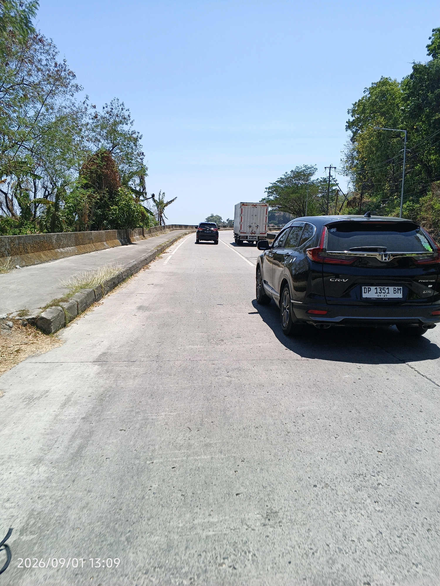 A black Honda CR-V, license plate DP 1351 BM, is traveling on a paved road. The vehicle is the most prominent subject in the foreground, its rear facing the camera. The road appears to be a highway or main thoroughfare, with multiple lanes visible. In the background, a white truck is also visible, suggesting ongoing traffic. The environment is characterized by a clear blue sky and lush green trees lining the sides of the road, indicating a possibly rural or semi-urban setting within Kupa, Indonesia. A pedestrian walkway with a concrete barrier is situated to the left of the road. The time stamp "2026/09/01 13:09" is superimposed on the lower left corner of the image.