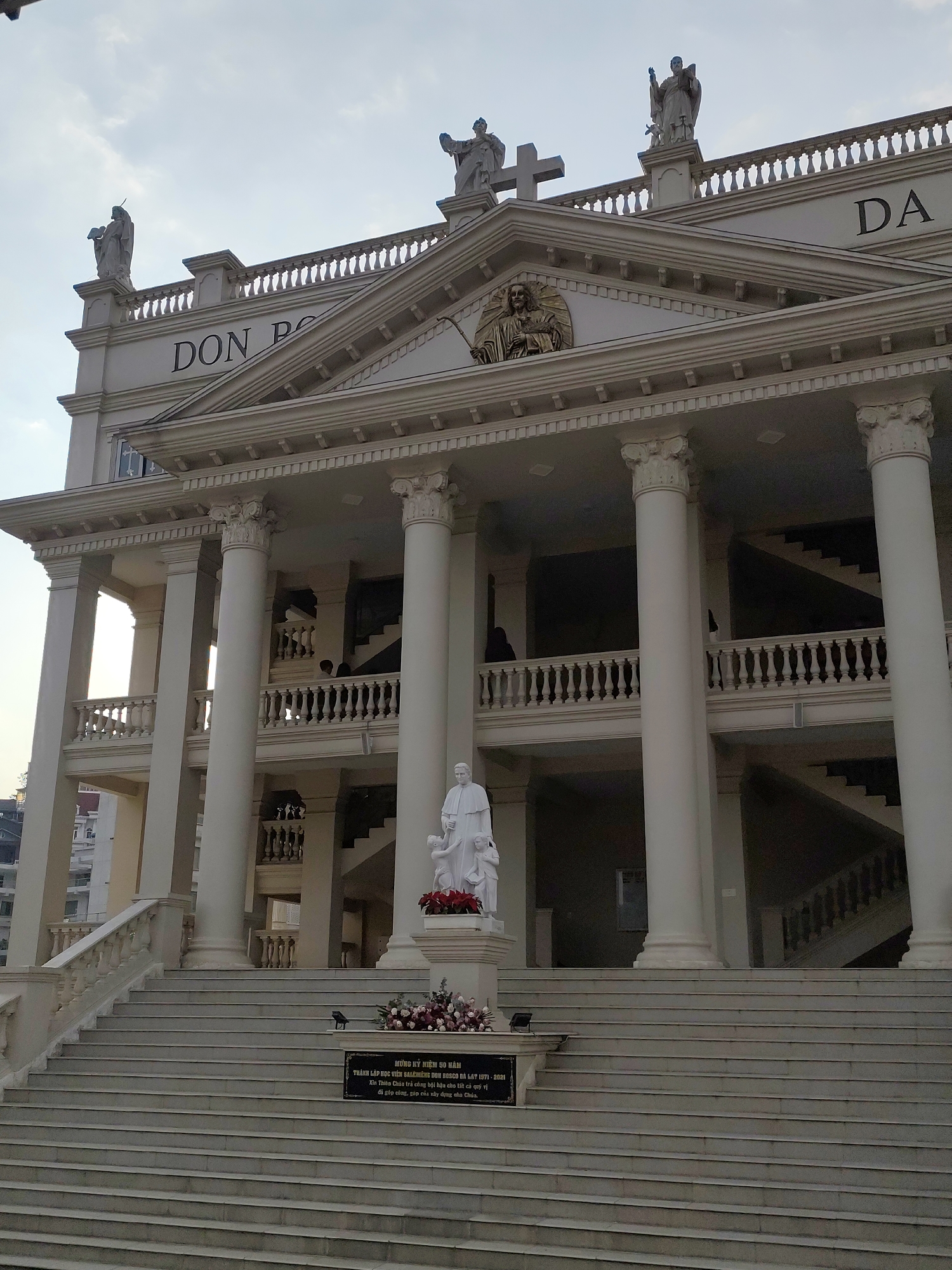 This image shows the Don Bosco School in Da Lat, Vietnam. The facade of the building is white with tall columns and balustrades. At the top, the name "DON PO" is visible on the left and "DA" on the right. There are statues on the roofline. A statue of a religious figure with children stands at the base of a long staircase leading to the building's entrance. A plaque with text in Vietnamese commemorating the 50th anniversary of the Salerian Don Bosco School is at the base of the statue. A building can be seen to the left, outside. The sky is overcast. Some individuals are visible in the background.