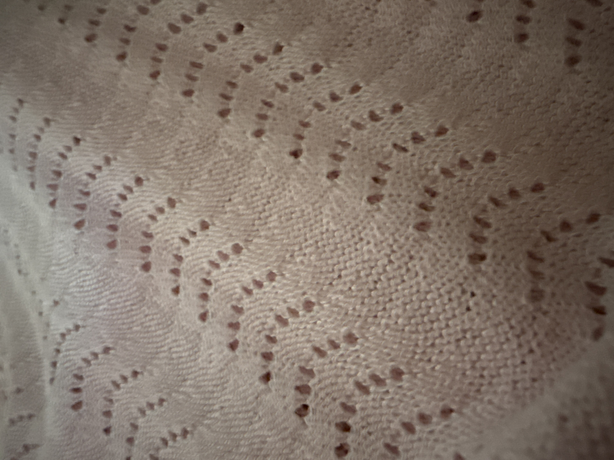 The image shows a close-up of a pink knitted fabric with a lace-like pattern. The pattern consists of small holes arranged in a zig-zag or wave-like design across the textile. The texture of the knit is visible, with individual stitches appearing as raised lines. The lighting casts subtle shadows, highlighting the dimensional quality of the pattern. The fabric appears soft and possibly delicate. There are no people, actions, or specific environmental elements visible in this close-up shot of the textile. The image is devoid of any context related to the city of Okhtyrka, Ukraine, or any other geographical location.