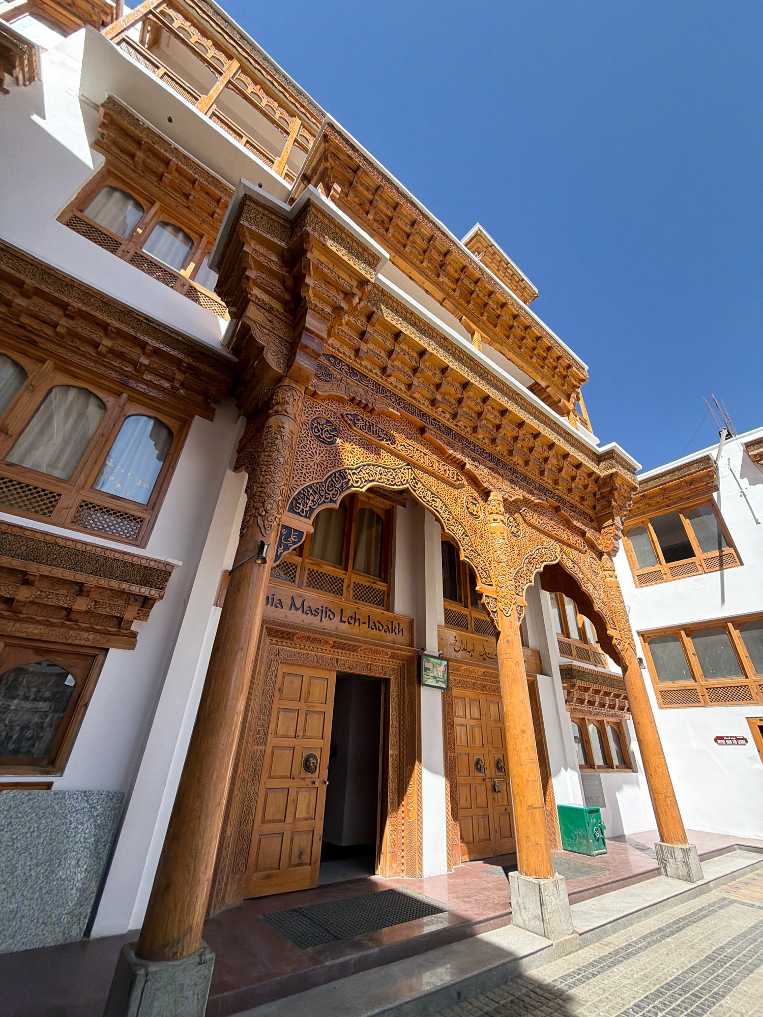 The image shows a low-angle, wide shot of the Jama Masjid in Leh, India. The mosque features intricate wooden carvings on its facade, balconies, and entrance archways. The building has white walls and is illuminated by bright sunlight. The entrance doors are made of wood with square panels and are partially open, revealing a dark interior. A sign above the entrance reads "Jama Masjid Leh-Ladakh". Green bins are visible near the entrance. The ground in front of the mosque is paved with stone tiles. The sky is clear and blue. The perspective is distorted due to the wide-angle lens and the upward tilt of the camera.