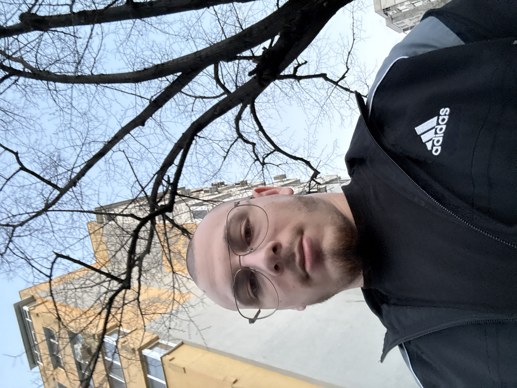 A man wearing an Adidas jacket and glasses is standing outside, facing the camera. In the background are bare tree branches, a clear sky, and buildings. Based on the architecture, the location may be in the city of Stara Zagora, Bulgaria.