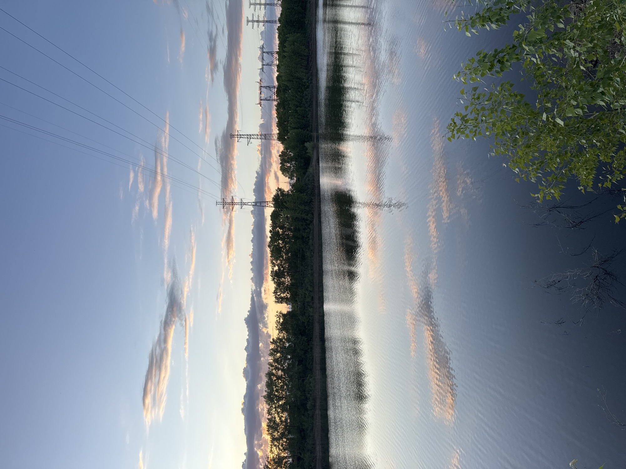 This media file captures a tranquil landscape scene in Chaikovsky, Russia, during what appears to be either sunrise or sunset.
The primary subjects are a large body of water, a line of trees, power transmission towers, and the sky. The image features a wide expanse of calm water in the foreground and midground, reflecting the colors of the sky and clouds. A dense, dark green treeline runs horizontally across the middle of the frame, separating the water from the distant horizon. Behind and above the treeline, a series of tall, metallic power transmission towers (pylons) are visible, extending into the distance. Multiple power lines stretch horizontally across the sky between these towers. In the immediate foreground on the right, vibrant green foliage from a tree branches into the frame, partially obscuring the water.
The sky is a clear gradient of blue, transitioning to warmer, lighter tones closer to the horizon. Scattered, elongated clouds appear to be lit by the low sun, exhibiting soft orange and pink hues, which are also beautifully reflected in the water's surface. The water is relatively smooth with subtle ripples, causing slight distortions in the reflections.
No specific activity or event is taking place; it is a static landscape view. The time of day is notable for its soft, warm lighting, indicative of either early morning or late evening. The scene evokes a sense of peace and vastness, highlighting a blend of natural beauty and industrial infrastructure. There is no visible text in the image.