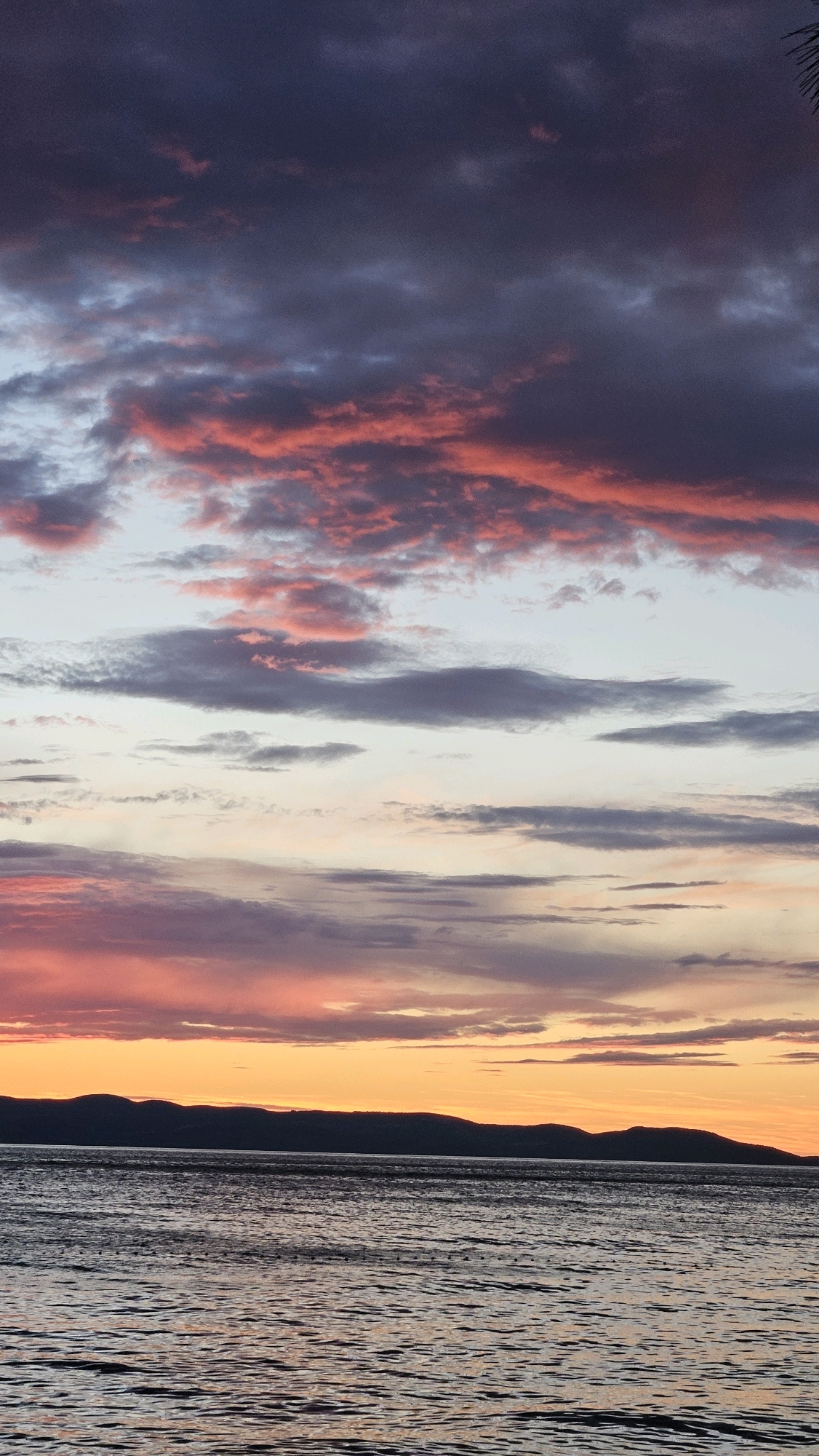 The image depicts a sunset over a body of water, likely the Adriatic Sea, viewed from the coast of Općina Tučepi, Croatia. The sky is filled with dramatic clouds, ranging in color from deep purple and gray to vibrant pinks and oranges, indicating the sun is low on the horizon. A dark, silhouetted mountain range forms a line across the distance, separating the sea from the sky. The water in the foreground is rippled, reflecting the colors of the sunset. There are no people or man-made structures visible in the scene.