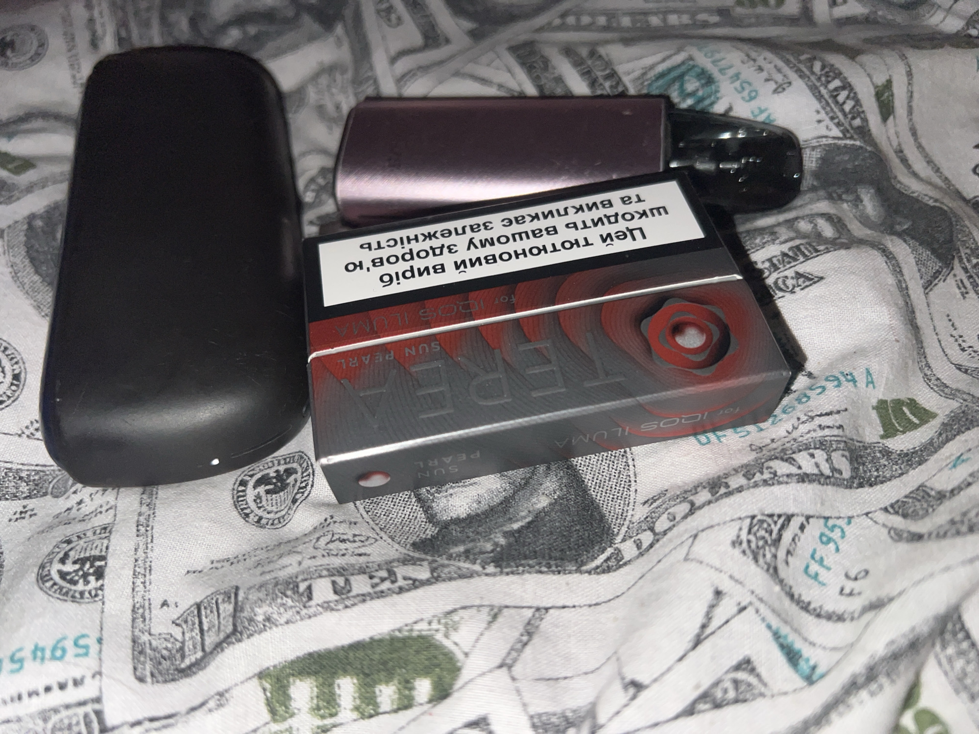 This is a close-up, overhead shot of a pack of IQOS ILUMA "SUN PEARL" HEETS, a rose-gold IQOS device, and a black IQOS holder, all resting on a surface covered with US dollar bills. The HEETS pack is open and partially obscured by the rose-gold device. The black holder is to the left of the HEETS pack. The rose-gold device is on top of the HEETS pack, partially covering it. The background is a white fabric printed with a pattern of US dollar bills, some of which are blurred. The image does not provide any information about the city of Zarudyntsi, Ukraine.