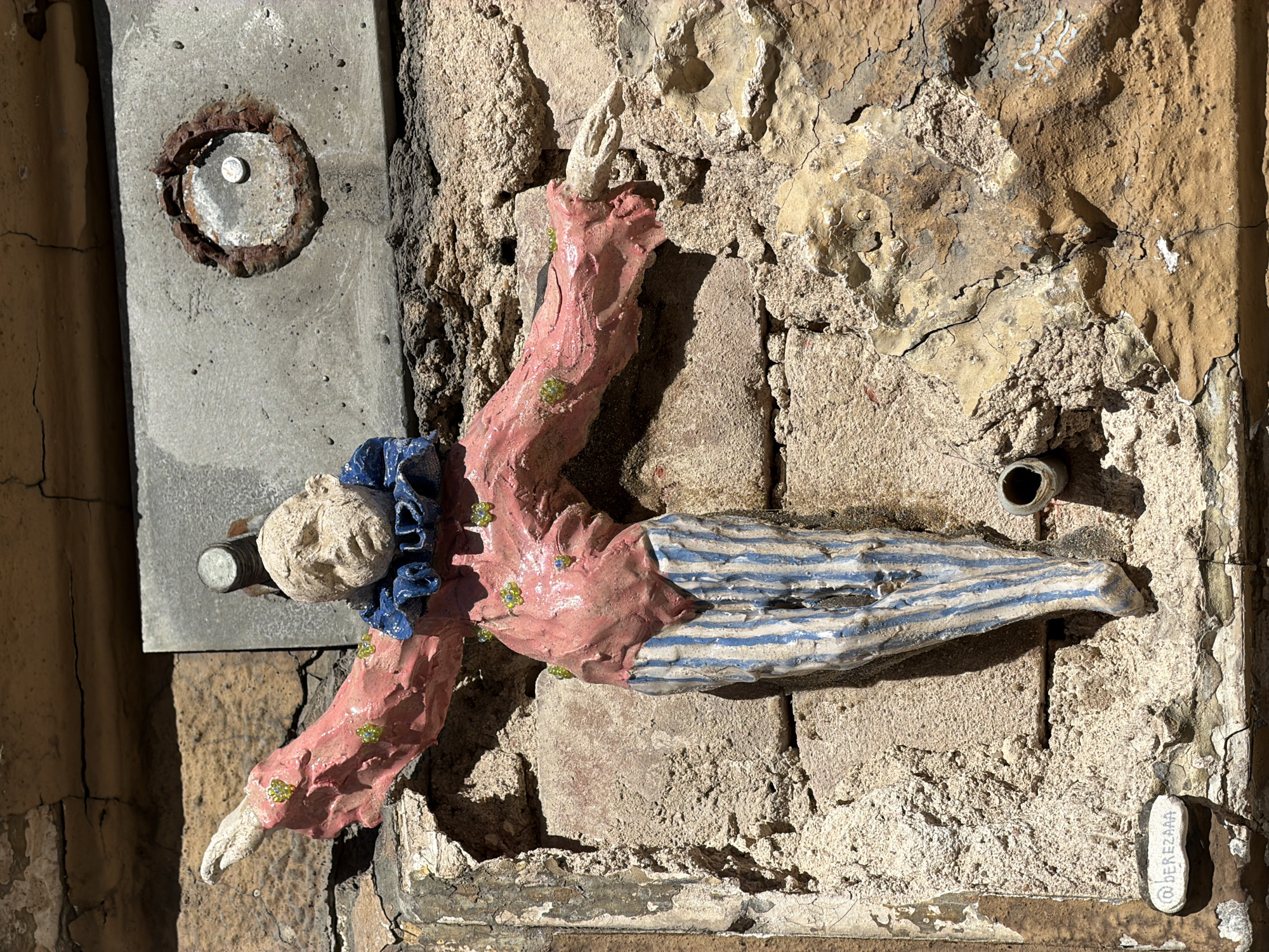 This image captures a weathered ceramic or plaster figure mounted on a distressed brick wall, characteristic of street art installations by the artist Maria Bereza, commonly found in Saint Petersburg, Russia. The figure, possibly depicting a melancholic clown or a stylized human, features a bald head with closed eyes and a somber expression, accentuated by a ruffled blue collar. Its torso and arms are painted a textured pink, adorned with small, irregular green and blue bead-like embellishments. The arms are outstretched horizontally, with open hands. The lower half consists of legs clad in vertically striped blue and white trousers.
The figure is affixed to an aged wall constructed of bricks, heavily covered in peeling, cracked, and discolored plaster in shades of beige, brown, and off-white, showing significant signs of decay and exposure. Above the figure's head, a rectangular grey panel, possibly concrete or metal, is visible, featuring a prominent rusted circular fitting with a small silver cap or bolt in its center, and another larger bolt or pipe extending upwards behind the figure's head. Below the figure's right leg, a small, open pipe or conduit is embedded in the wall. In the lower left corner, a small, light-colored rectangular plaque is mounted, bearing the inscription "@BEREZAAA". The lighting suggests direct sunlight, creating distinct shadows and highlighting the rough textures of both the figure and the wall.