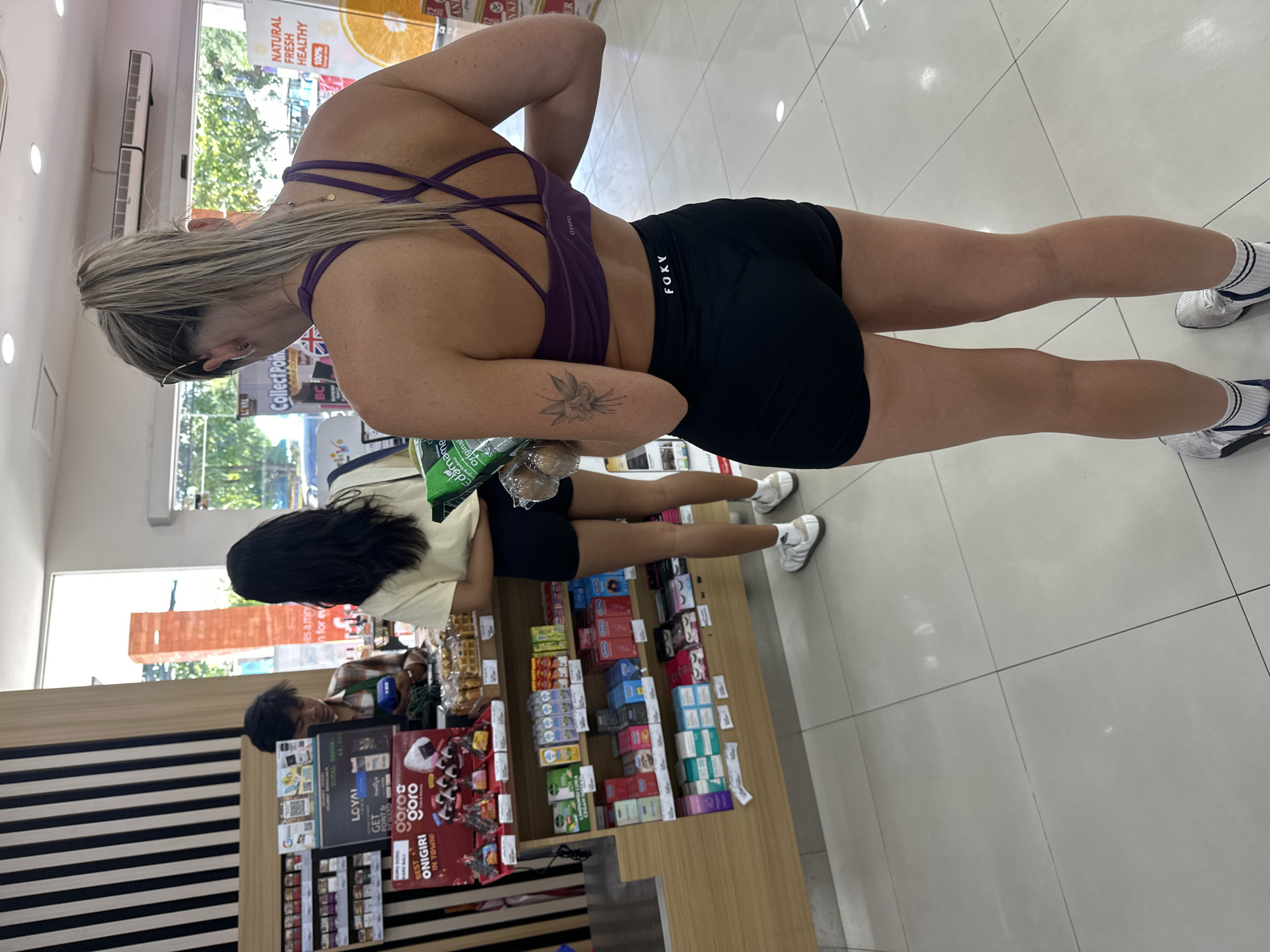 The image depicts the interior of a convenience store, likely a minimart, in North Kuta, Indonesia. The scene is captured from behind a woman wearing a purple sports bra and black athletic shorts, who is holding a green bag of what appears to be edamame and a clear plastic bag with items inside. She is standing in an aisle, facing a checkout counter where a male store employee in a plaid shirt is interacting with another customer. This customer, a woman with long dark hair, is wearing a light-colored t-shirt and dark shorts. The store shelves are stocked with various products, including packaged food items, health and beauty products, and beverages. The flooring is light-colored tile, and the lighting suggests an indoor environment. Natural light streams in from large windows at the back of the store, revealing greenery outside. The atmosphere is typical of a retail setting where customers are purchasing goods.