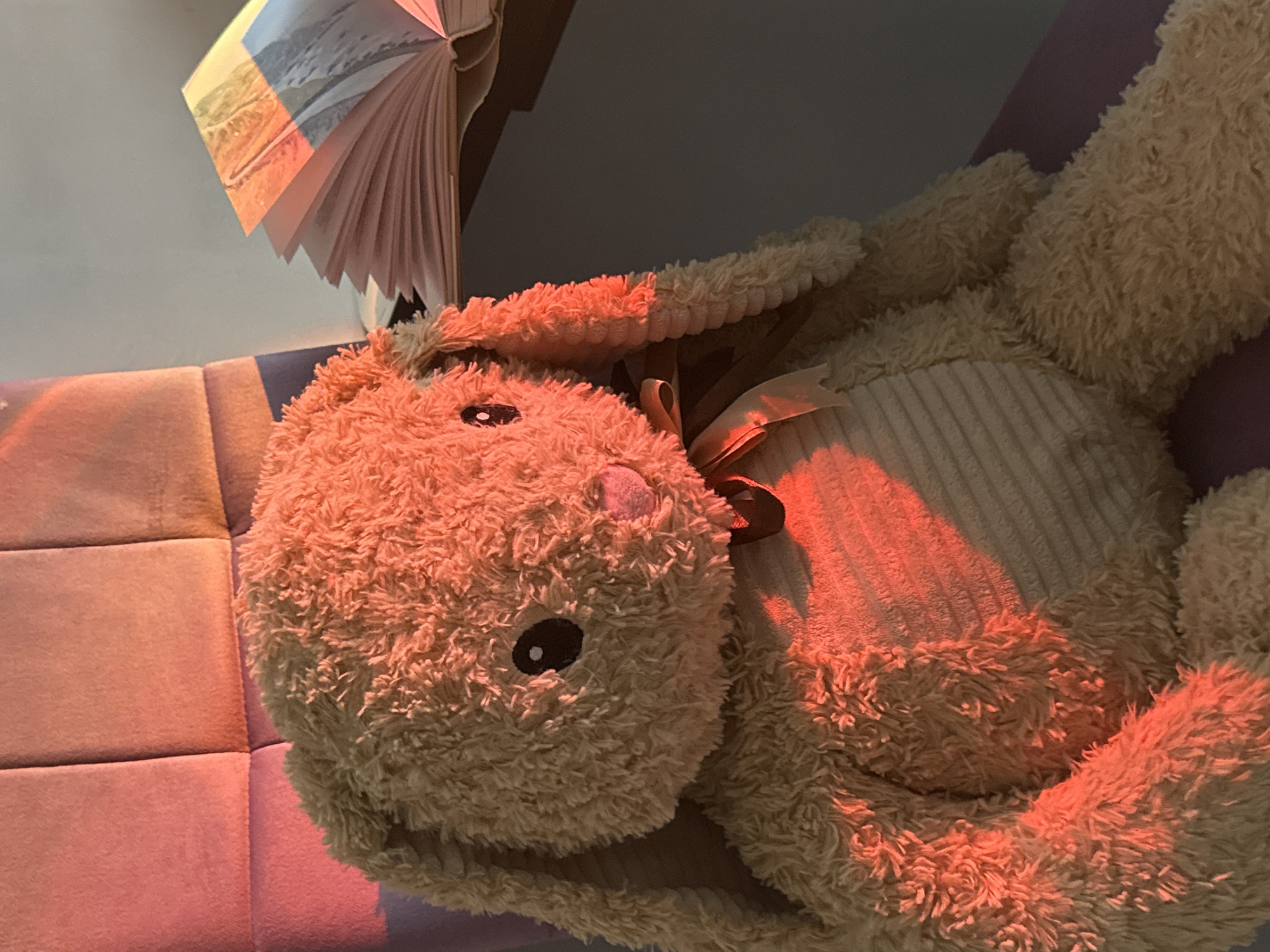 The image shows a stuffed bunny lying on a multi-colored upholstered surface. The bunny is light brown with a darker brown ribbon around its neck. The upholstery is patterned with squares of various shades of pink and purple. A book is partially visible in the upper left corner of the image. The lighting is warm and reddish. Location: Unknown, Unknown.