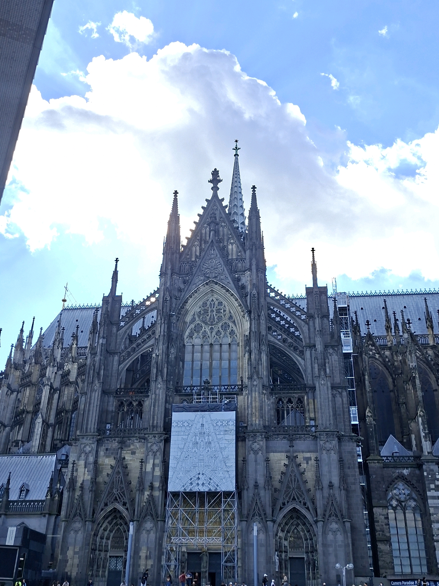 The image shows a low-angle, full-frame shot of the Cologne Cathedral (Kölner Dom) in Cologne, Germany, on a partly cloudy day. The gothic cathedral is a massive, dark stone structure with intricate architectural details, including pointed arches, spires, and ornate carvings. A large banner depicting a schematic of the cathedral is displayed on its facade, partially covering the windows and scaffolding. Sunlight pierces through the clouds, creating dramatic lighting effects on the building and sky. The foreground shows the base of the cathedral, with several people visible, some standing and others appearing to be walking. The environment is urban, with a modern building partially visible on the left edge of the frame, suggesting its proximity to other structures. The sky is a vibrant blue with scattered white clouds.