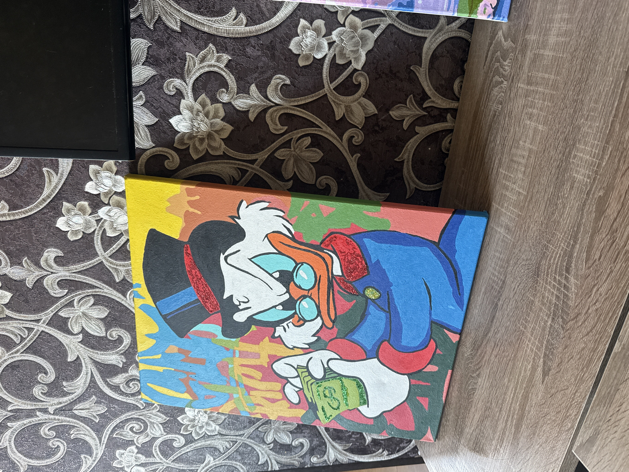 A close-up photograph shows a canvas painting depicting a cartoon character resembling Scrooge McDuck, with an orange beak, blue spectacles, a black top hat, and a blue coat with red trim, holding a stack of green bills in his white gloved hand. The artwork is brightly colored, featuring yellow, orange, red, blue, and green hues in an abstract manner. The painting is lying on a surface with brown and white floral wallpaper, and part of a wood-like grain surface is visible on the right side. A black rectangular object is seen to the left of the painting. The location appears to be indoors in an unknown city.