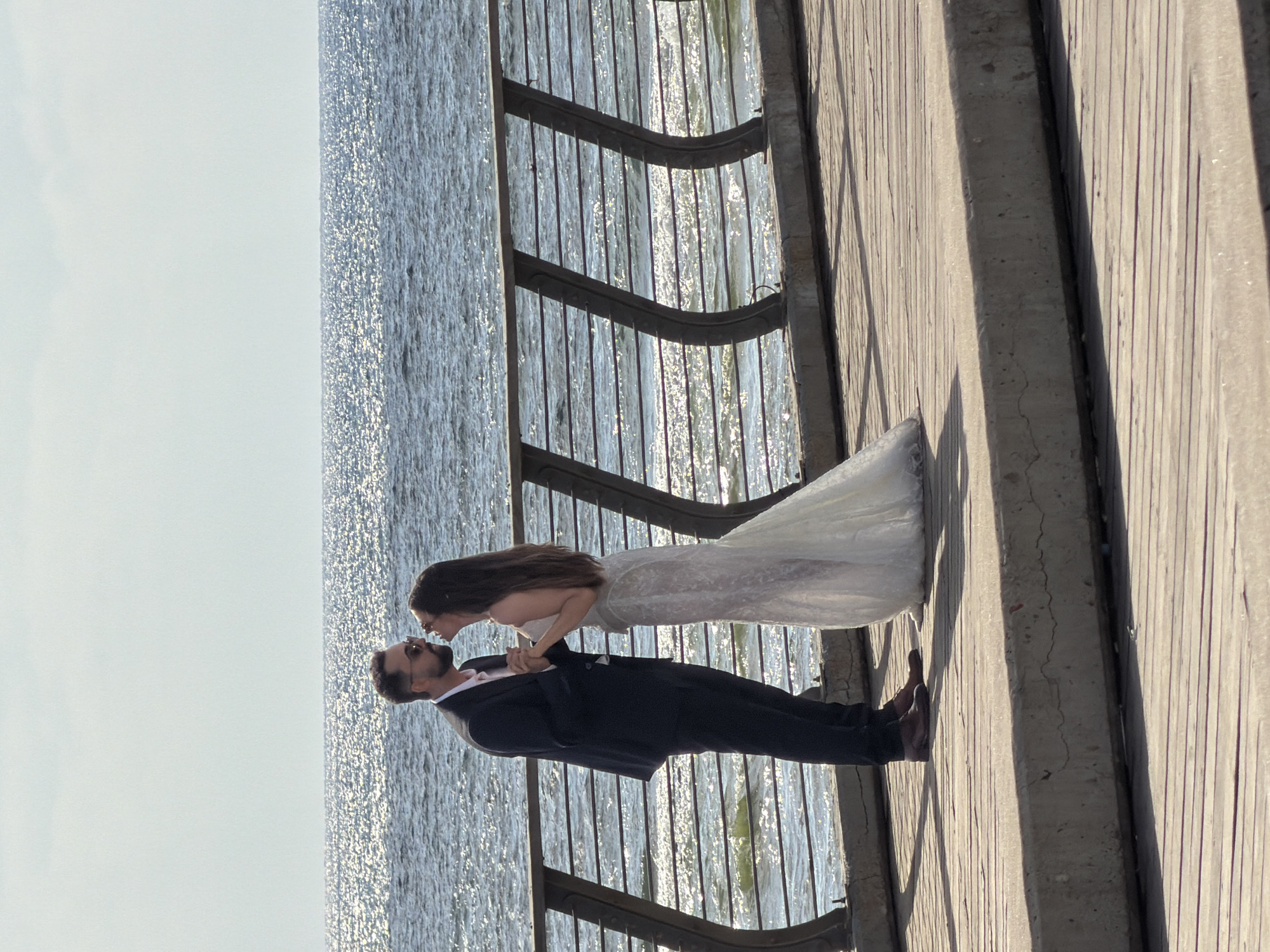 A newlywed couple is captured in a tender moment on a boardwalk by the sea. The bride, wearing a white lace wedding gown, leans in to kiss her groom, who is dressed in a sharp black suit and tie. Their faces are close, and their hands are clasped, suggesting a deep connection and affection.
The scene is set against the backdrop of the Mediterranean Sea, its surface shimmering with sunlight. A weathered wooden boardwalk, likely part of a promenade or pier, stretches out beneath them, bordered by a dark metal railing. The light indicates it's a bright, clear day, possibly late afternoon or early evening given the long shadows cast on the boardwalk. The location context points to Tel Aviv, Israel, a city known for its beautiful coastline and vibrant boardwalks. The overall mood is romantic and intimate, a snapshot of a joyous celebration. There is no visible text in the image.